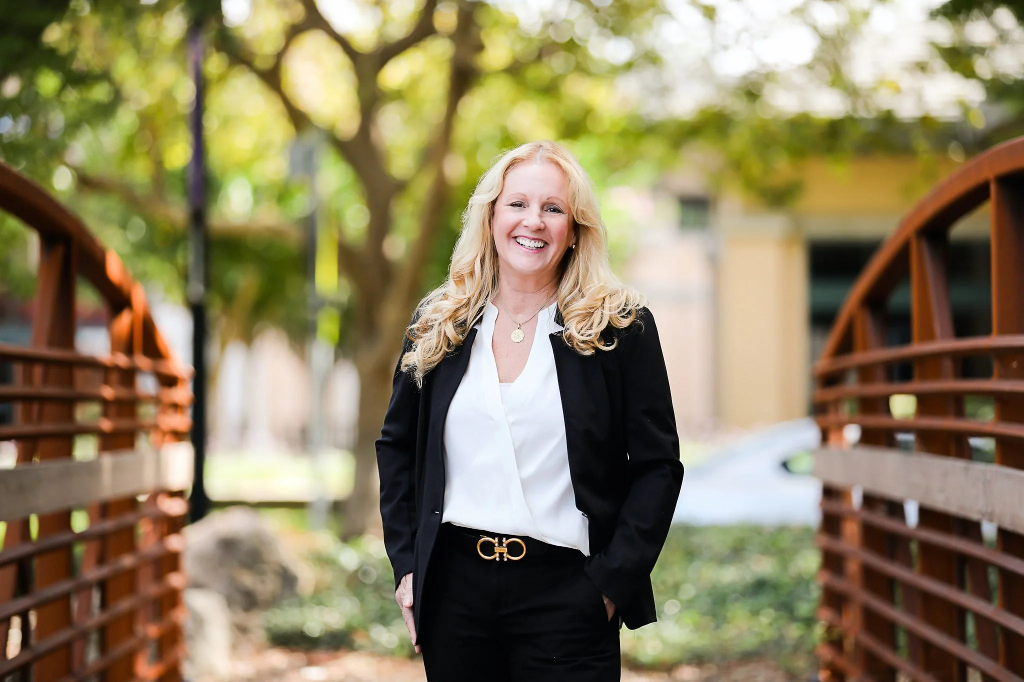 A smiling woman in business attire walking outdoors, with trees and a wooden bridge in the background.
