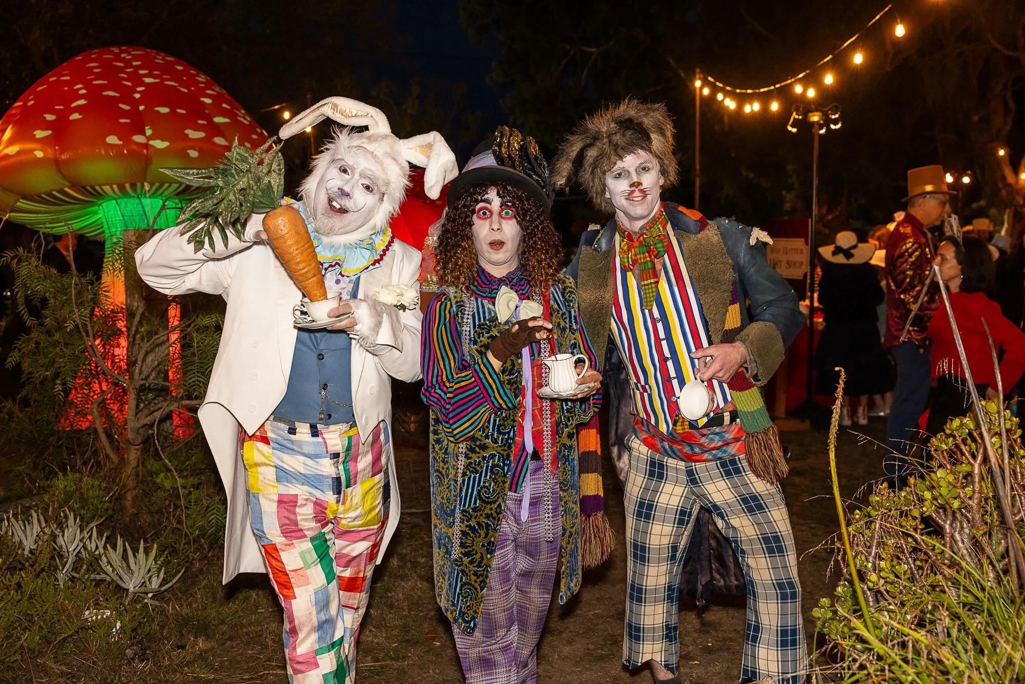 People dressed as colorful, whimsical clowns and characters at a party or event with string lights overhead.