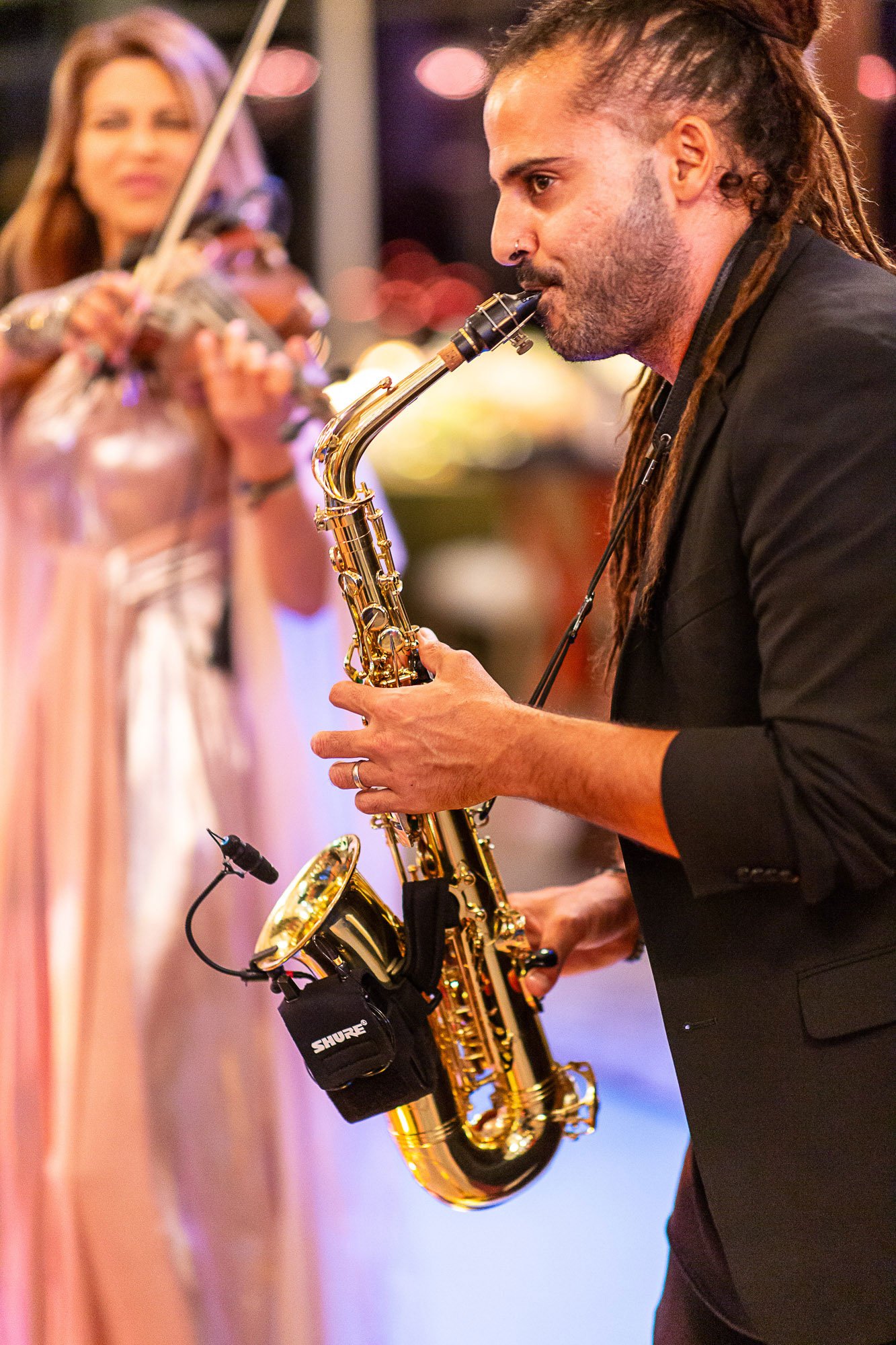 A man with dreadlocks playing a saxophone with a woman in the background playing a violin at an indoor event with colorful lighting at the Beach and Tennis Club in Pebble Beach, CA.