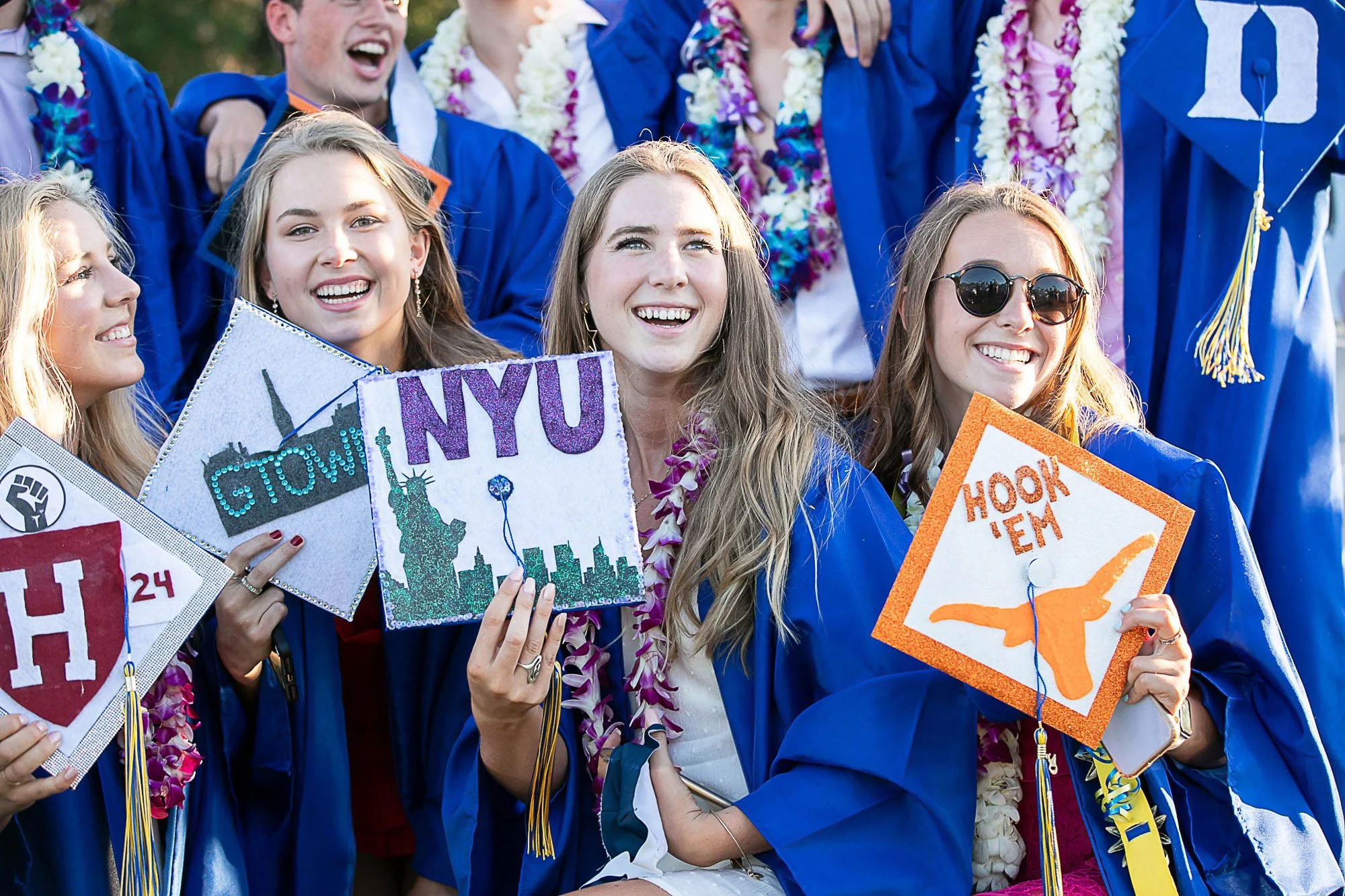 Group of graduates in blue caps and gowns celebrating outdoors, wearing flower leis, holding decorated signs and smiling.