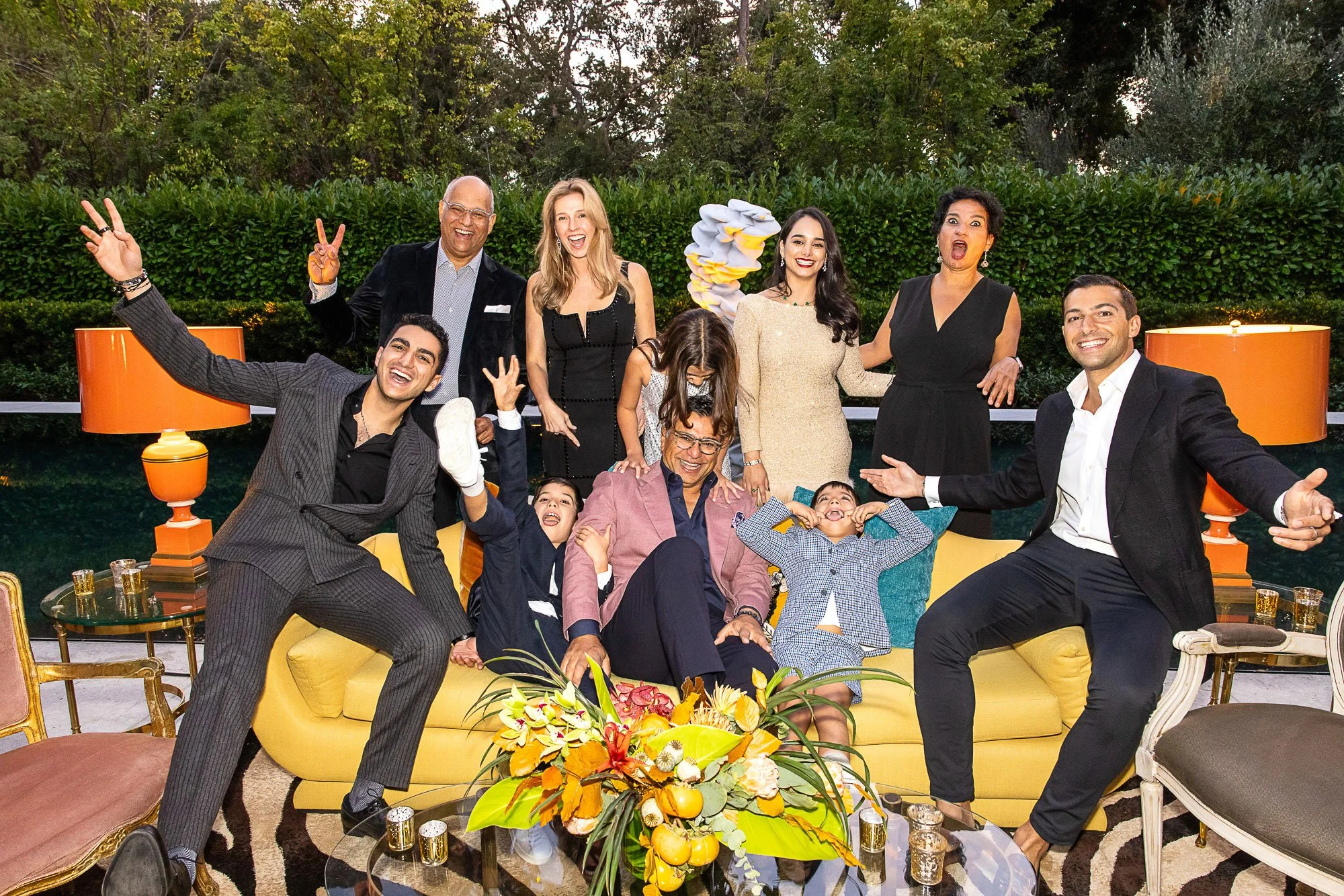 A diverse group of adults and children enjoying a gathering outdoors in the evening, smiling and posing happily, with a lush green hedge and trees in the background, decorated with orange lamps and a floral centerpiece.