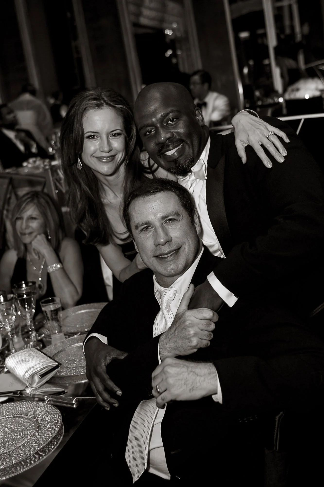 John Travolta dressed in formal attire at a social event, including a woman with long hair, a man with a beard and tuxedo, and a woman with glasses. They are sitting at a table with elegant dinnerware and glasses, smiling and posing for the camera.