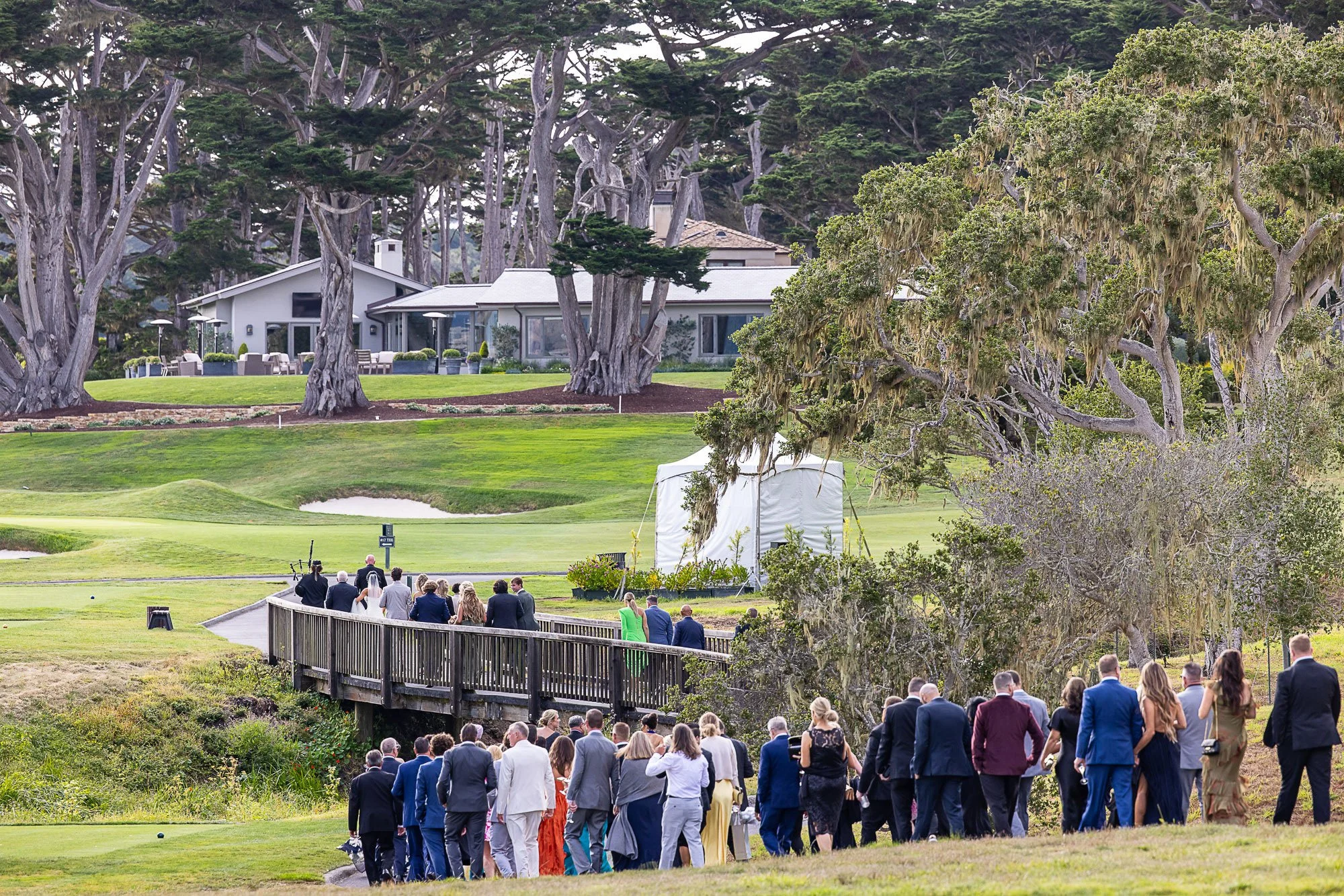 Wedding ceremony at the Beach and Tennis Club in Pebble Beach, CA outdoors on a golf course with guests walking on a pathway. Large trees and a modern house are in the background.