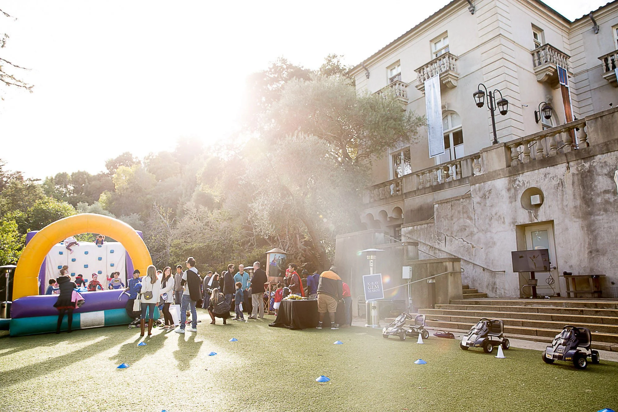 Outdoor event with children playing on an inflatable bounce house, surrounded by adults, in front of a historic building with classical architecture, trees, and sunlight.