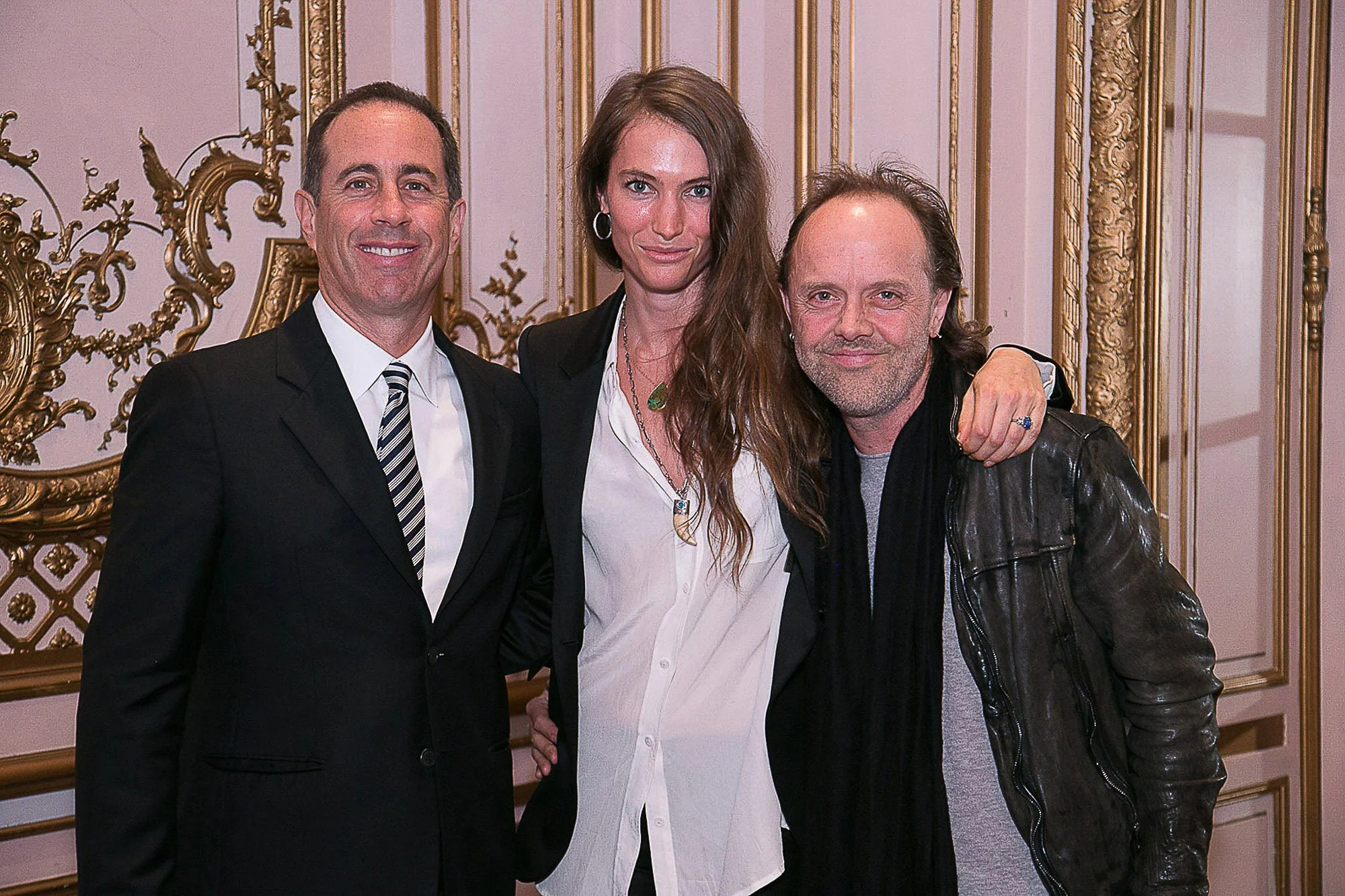 Jerry Seinfeld and two people pose together indoors, a man in a suit, a woman with long brown hair, and a man with long hair and a leather jacket, in front of a ornate gold decorative wall.