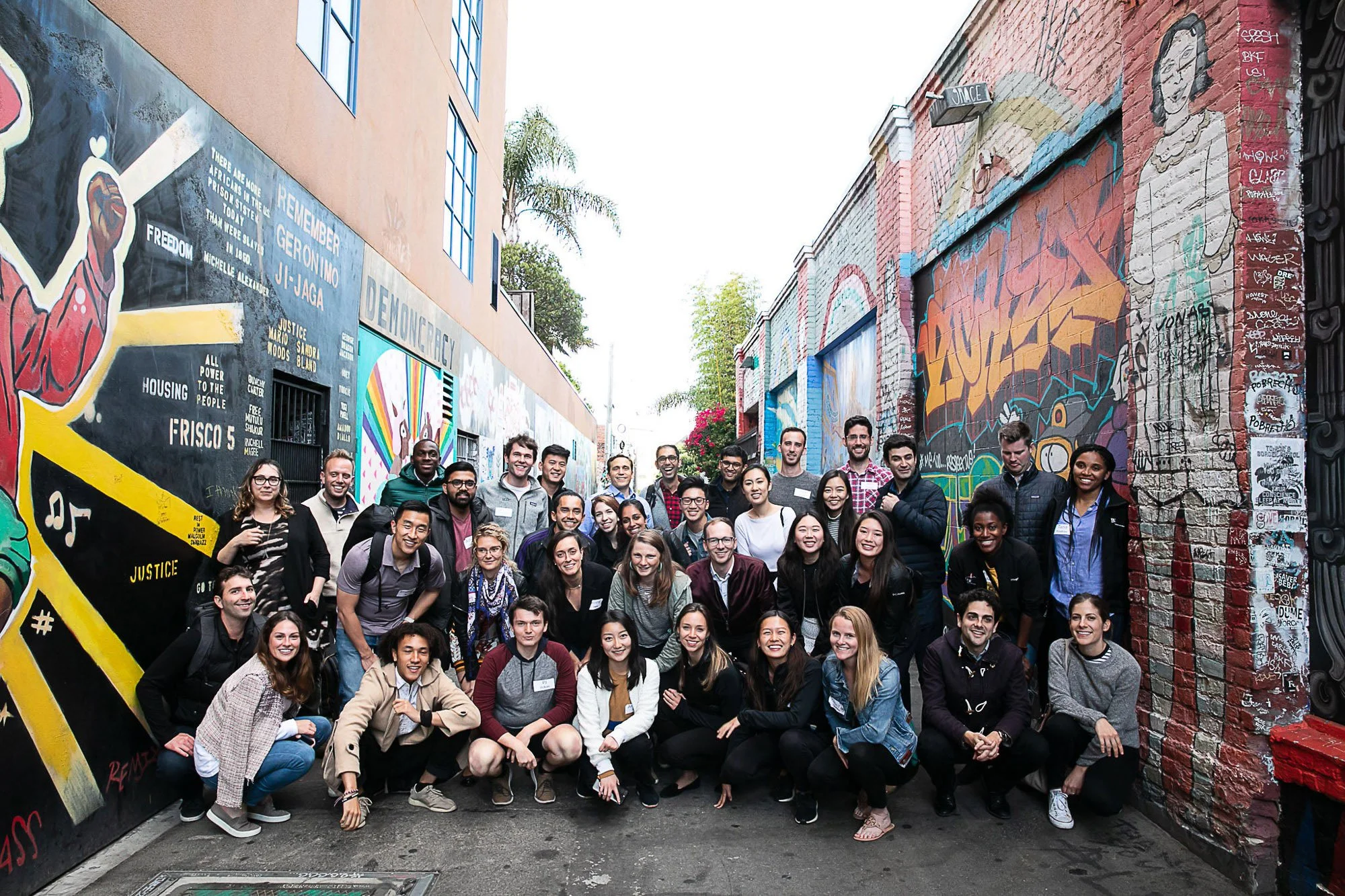 Large group of diverse people posing together in an alleyway covered with colorful murals and graffiti art.