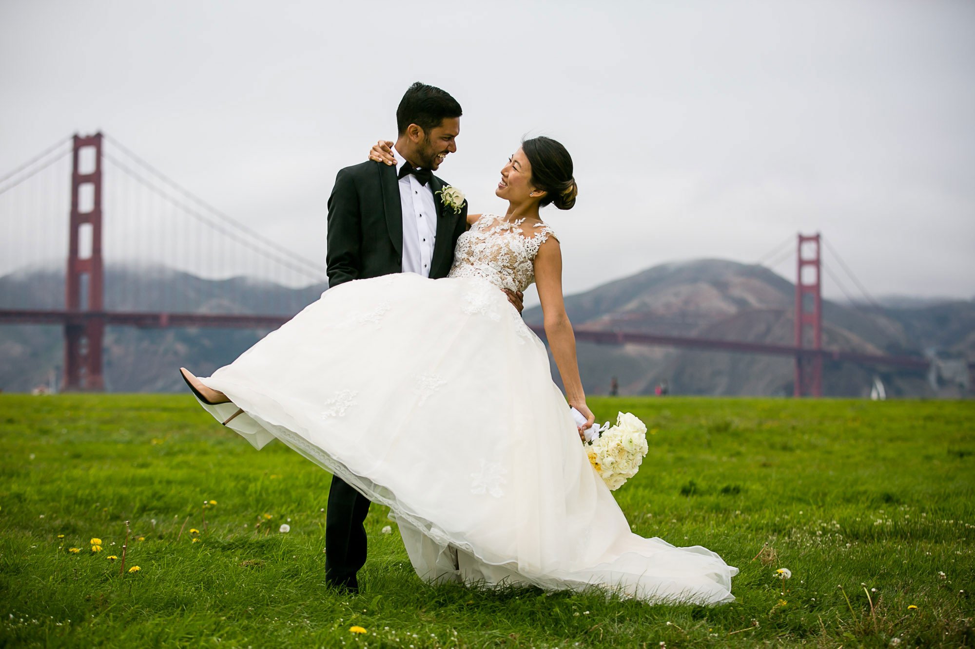 A newlywed couple, with the groom in a black tuxedo and the bride in a white lace wedding gown, standing on grass with the Golden Gate Bridge and hilly landscape in the background during overcast weather.