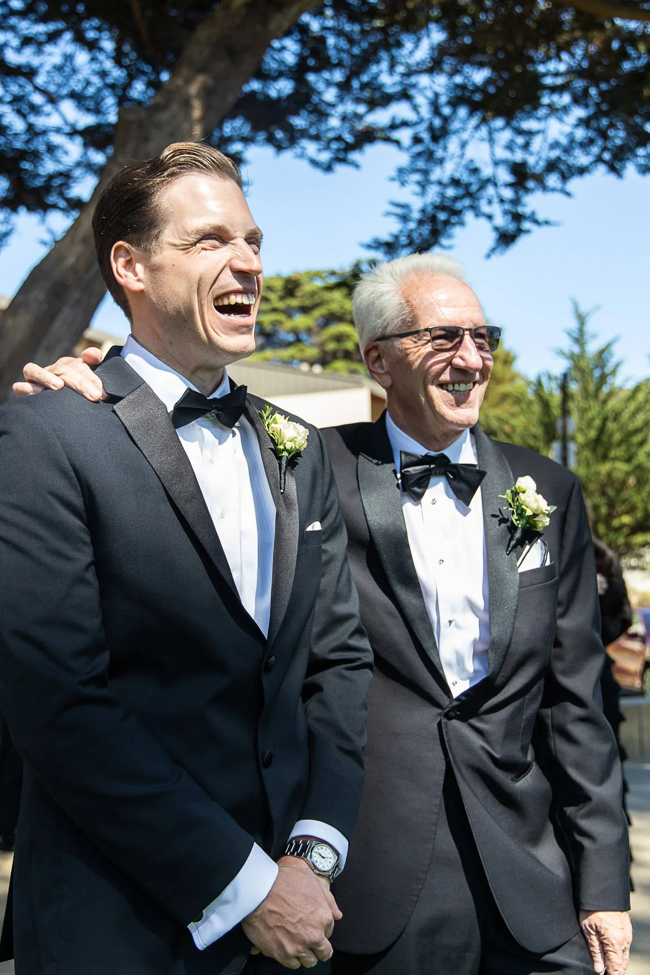 Two men dressed in tuxedos with boutonnières, standing outdoors under a large tree, smiling and enjoying each other's company at Cavallo Point in Sausalito, CA.