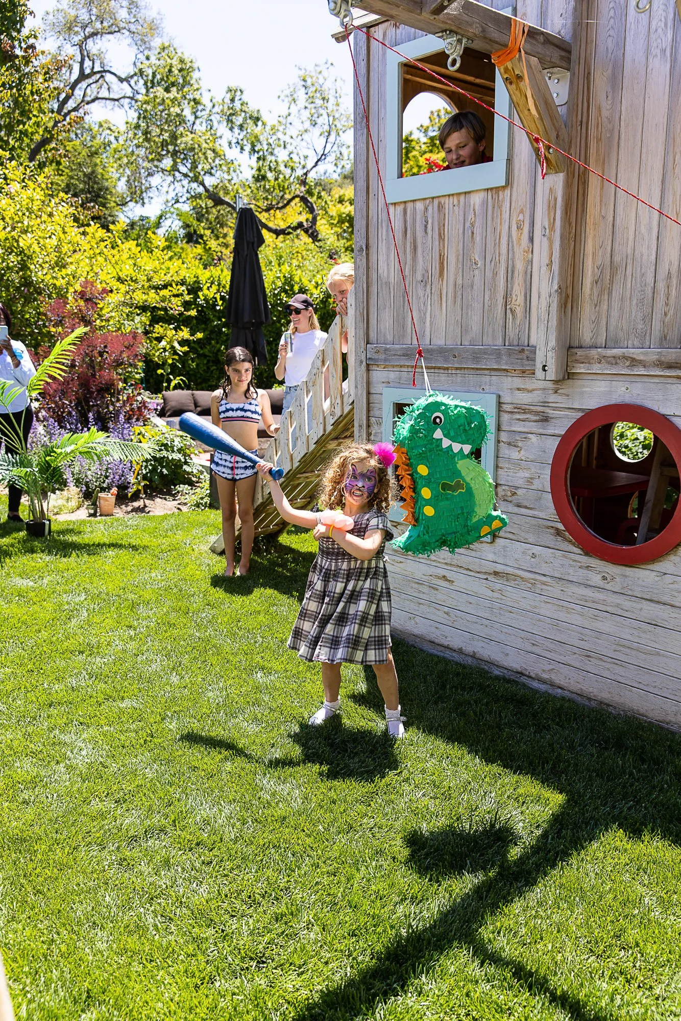 Children playing outside during a sunny day, with a wooden playhouse decorated with a dragon piñata hanging on the side, and people watching and taking photos in the background.