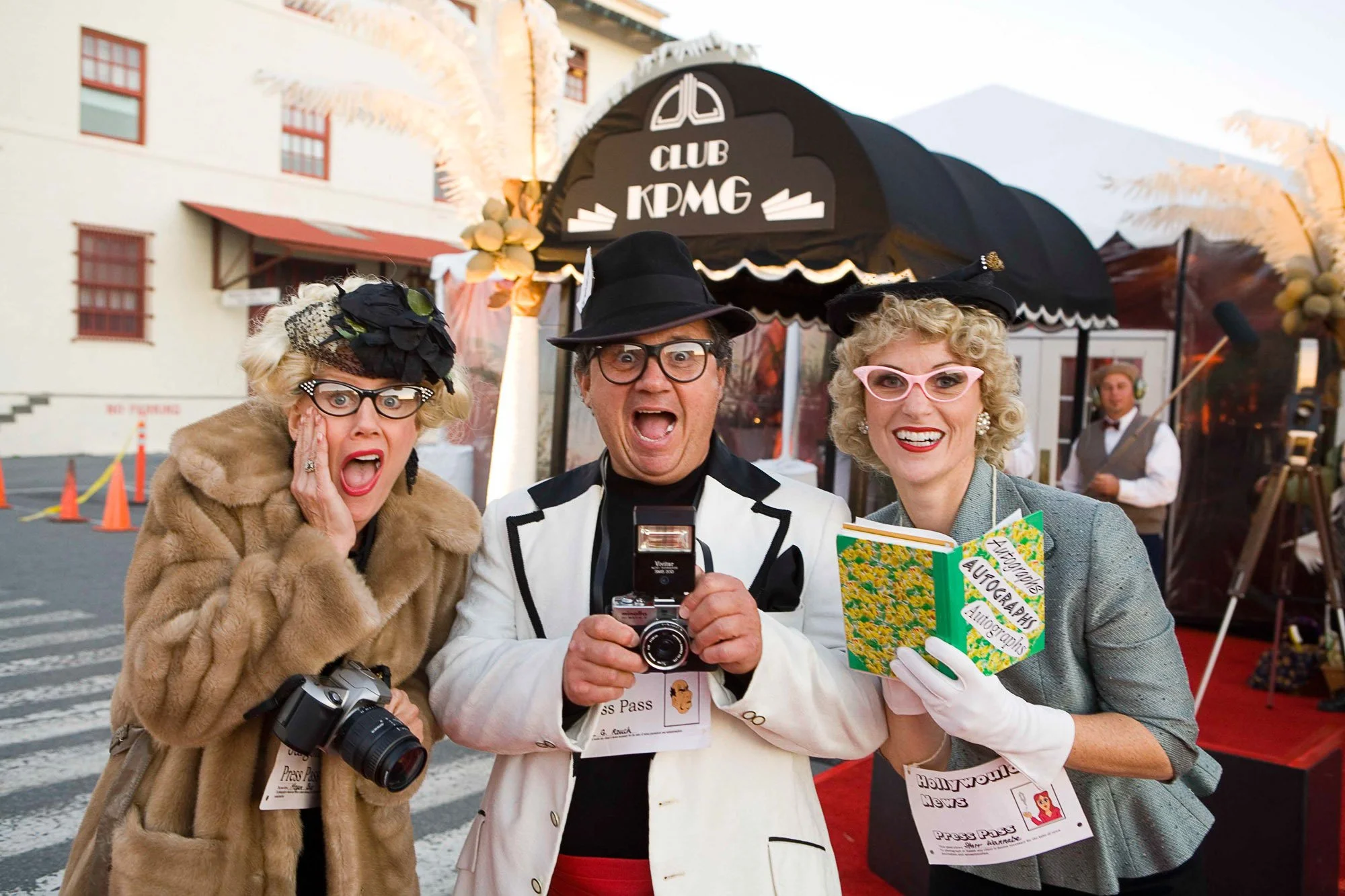 Three cheerful people dressed in vintage clothing, wearing glasses and hats, standing in front of a black tent with a "Club KPMG" sign, taking a selfie together at an outdoor event.
