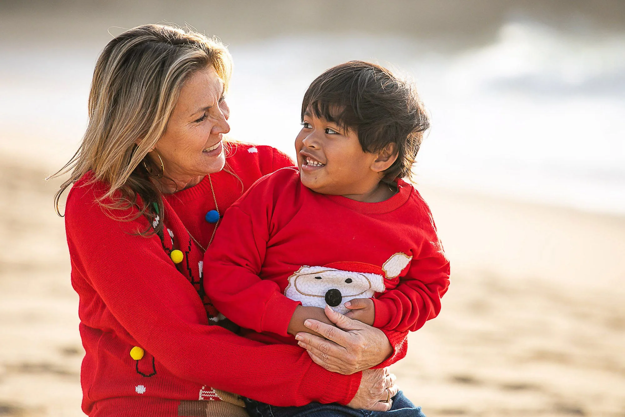 An elderly woman with blonde hair, wearing a red Christmas sweater with colorful pom-poms, holding a young boy with black hair, also wearing a red sweater with a dog design, at Montara Beach in Half Moon Bay.