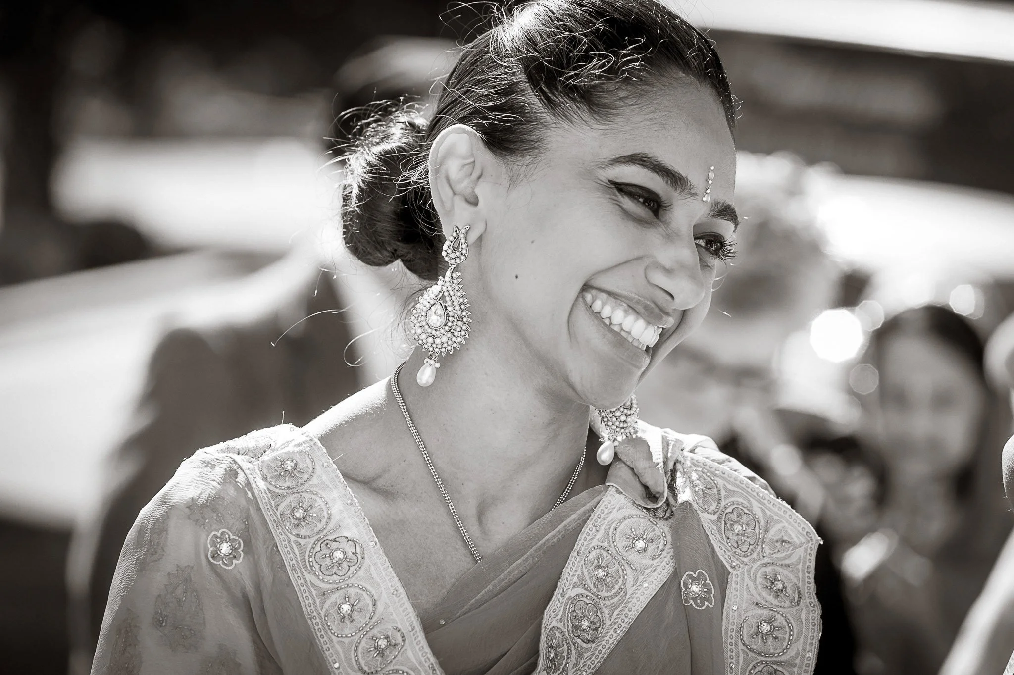 A smiling woman wearing traditional attire with intricate embroidery, large ornamental earrings, and forehead jewelry, standing outdoors with a blurred background.