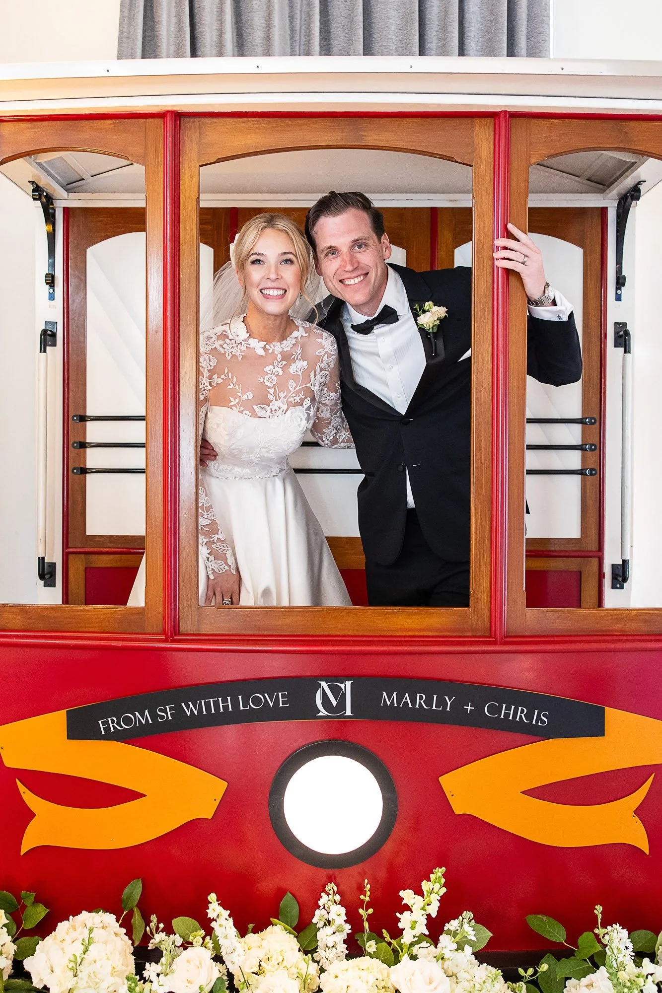 A bride and groom smiling inside a wooden trolley decorated with orange and yellow flames, with a sign that reads 'From SF with Love.' The bride wears a white lace wedding gown, and the groom is in a black tuxedo with a bow tie. There are white flowe