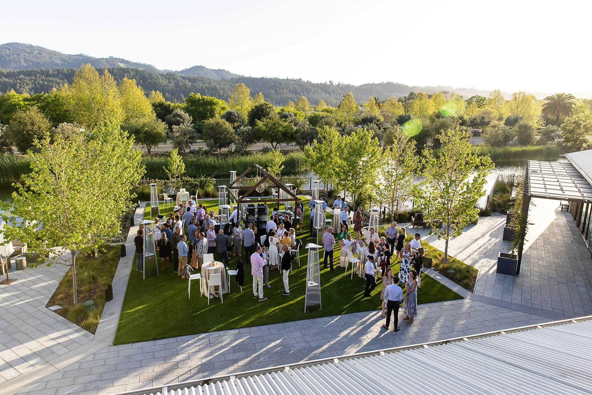 Outdoor gathering at Napa Valley Four Seasons with trees, mountains, and a body of water, where people are socializing around tables and standing on a green lawn during daylight.