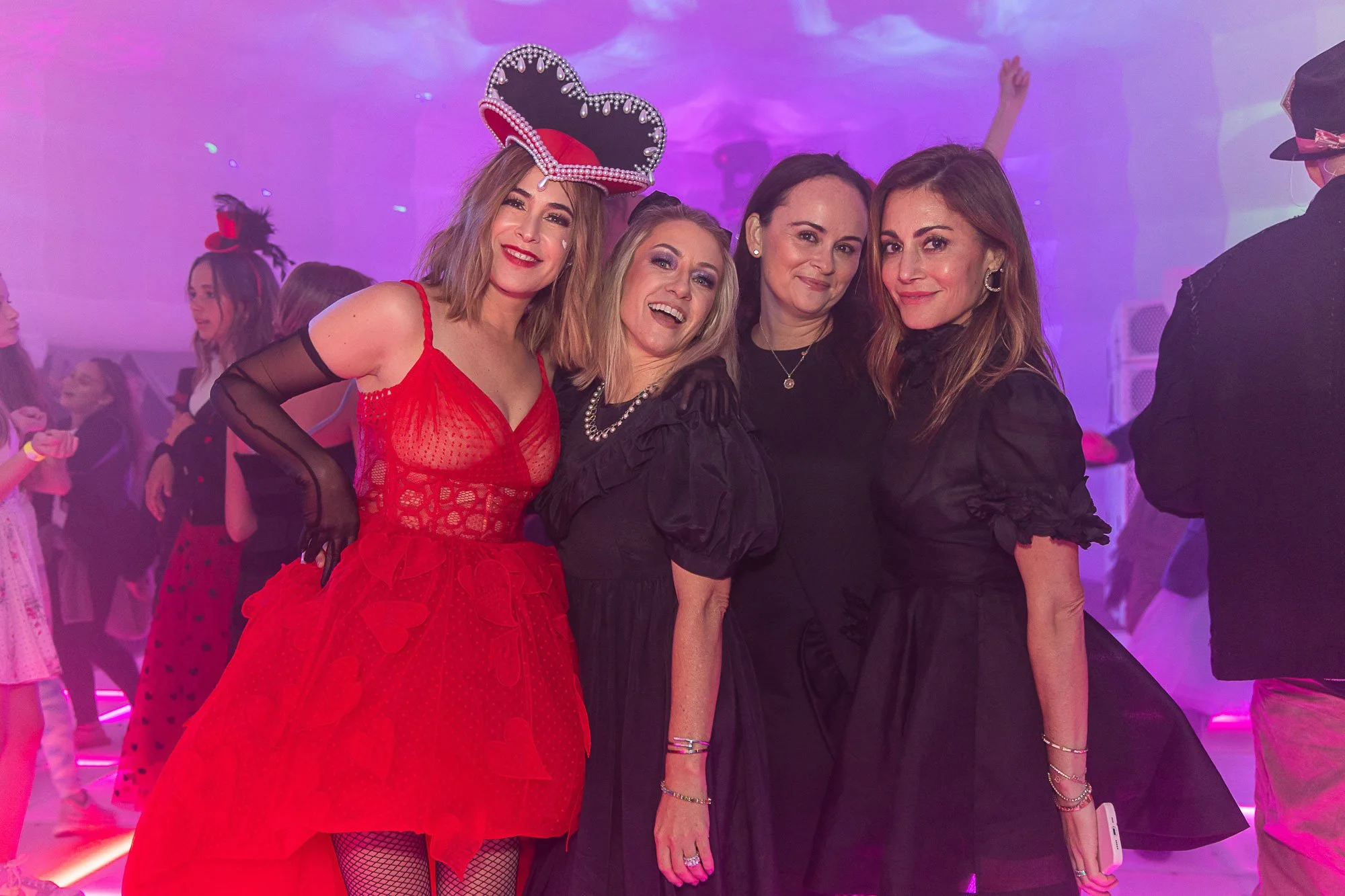 Four women smiling at a party with purple lighting. One woman wears a red dress and a heart-shaped crown. The women are dressed in festive attire, surrounded by other partygoers.