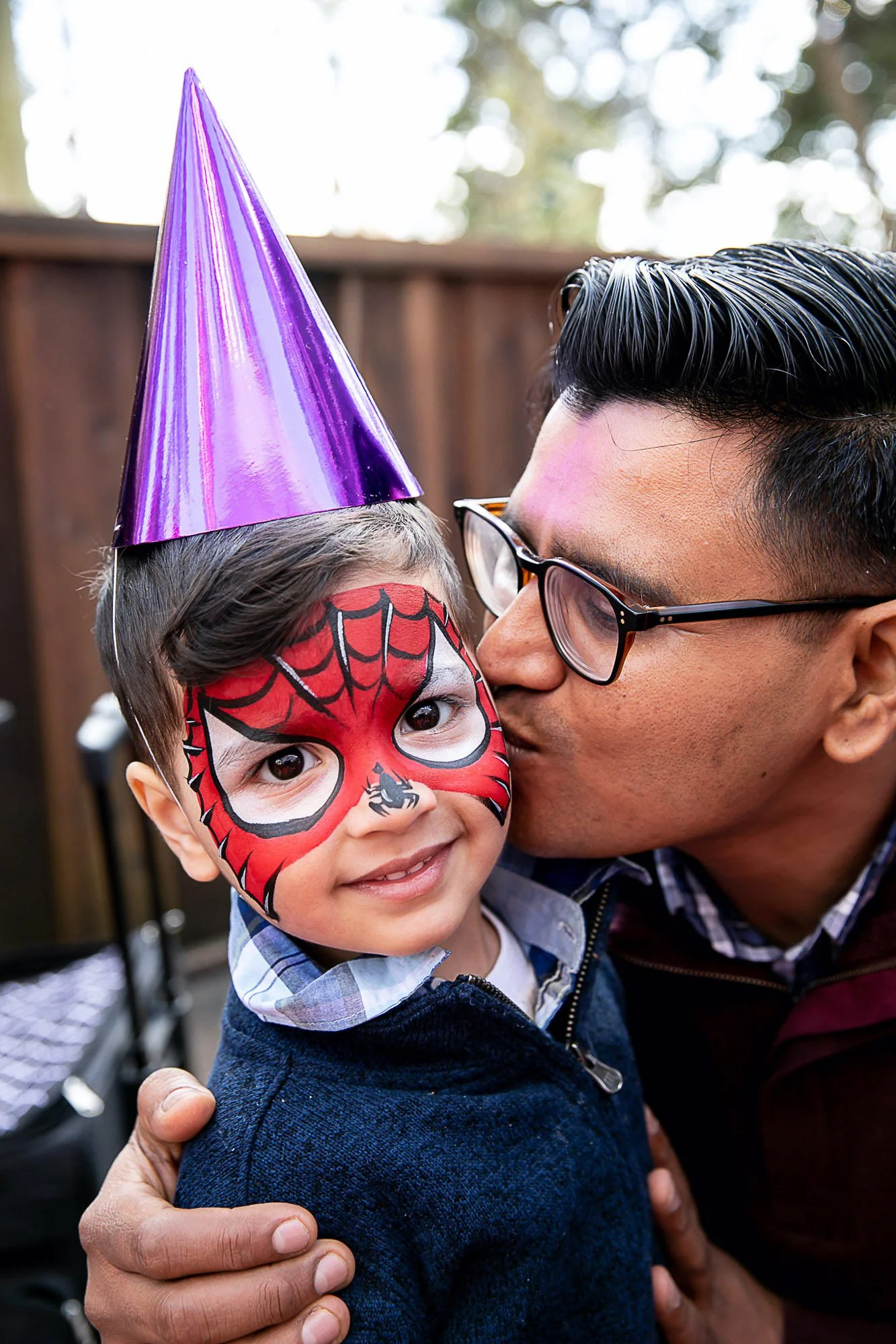 A young boy with face paint resembling Spider-Man and a purple party hat, smiling as an adult man leans in to kiss him on the cheek in an outdoor setting.
