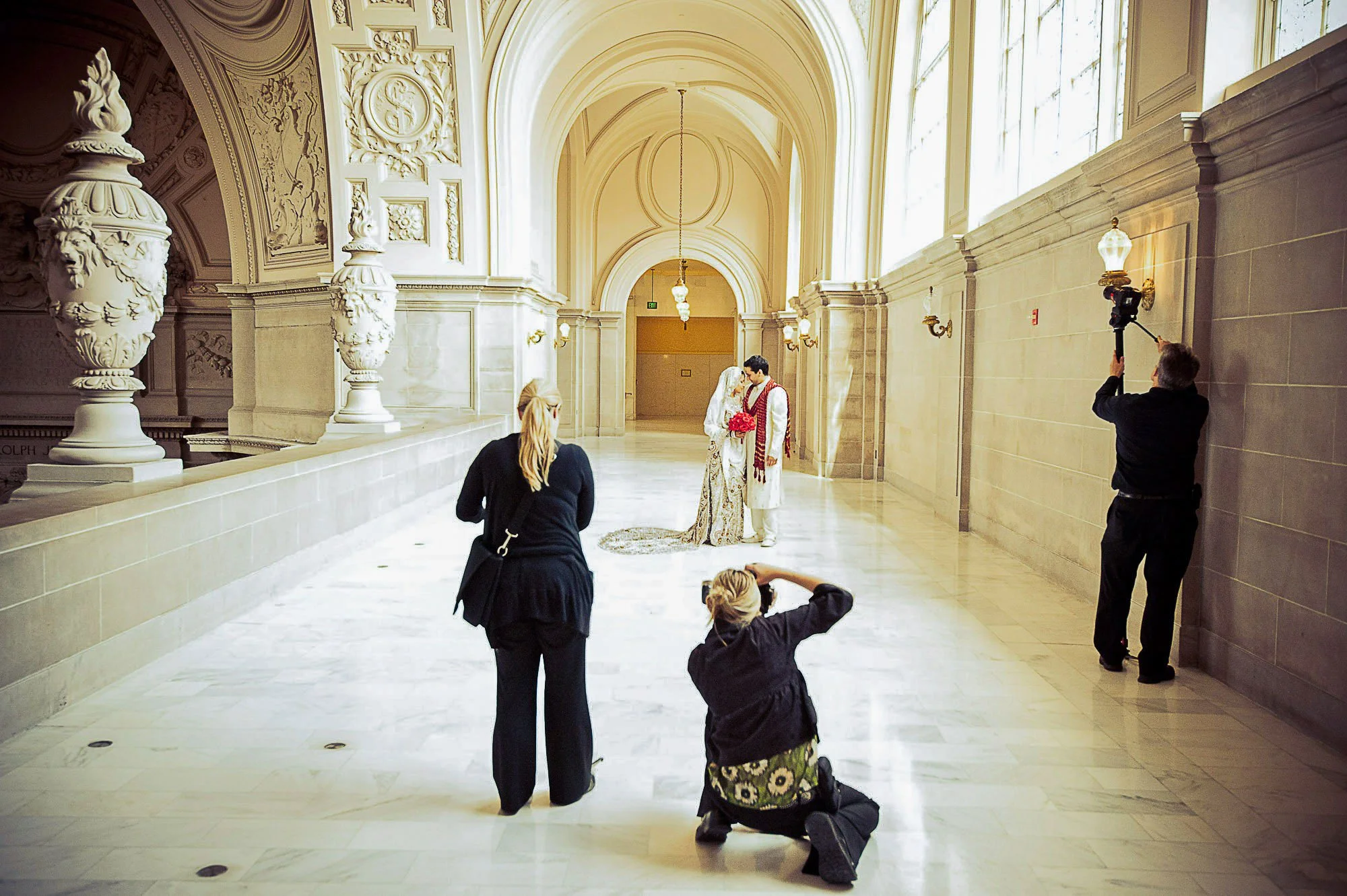 A wedding photoshoot taking place in a grand, elegant indoor hall with high arched ceilings and ornate wall details. Two people dressed in wedding attire are standing closely, while four other women and a man are positioned around them, photographing