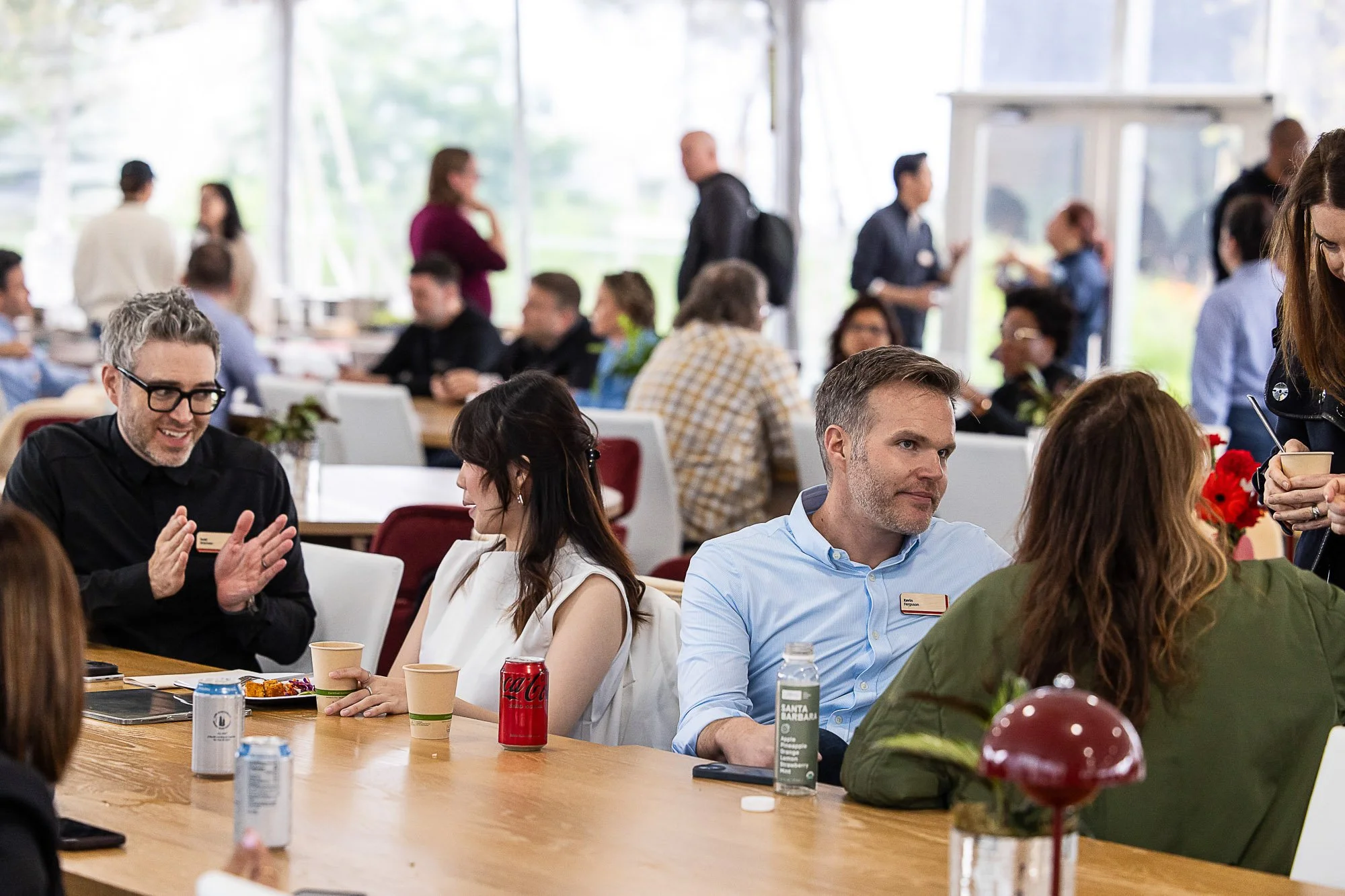 People sitting at a long table in a bright room, engaging in conversation, with some standing and chatting in the background, during a social gathering or event.