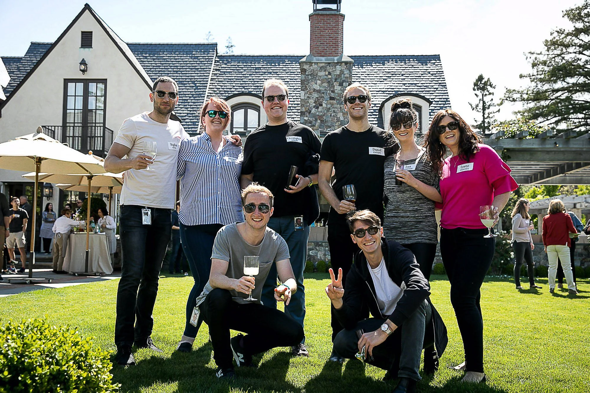 Group of six friends at a daytime outdoor gathering in front of a large house with stone chimney, holding wine glasses and smiling, sunny weather, some wearing sunglasses.