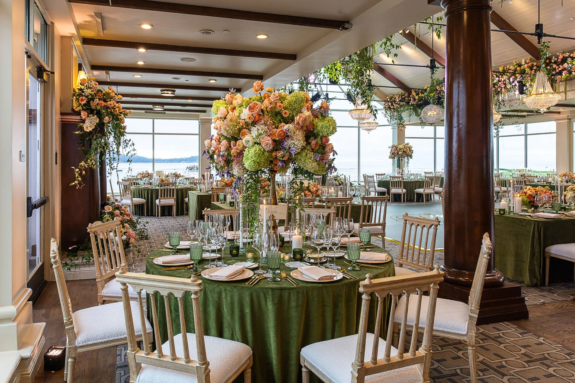 Elegant indoor event space decorated with large floral centerpieces, with round tables set with white napkins, glassware, and green cups, and overlooking a waterfront view through large windows at the Beach and Tennis Club in Pebble Beach, CA.