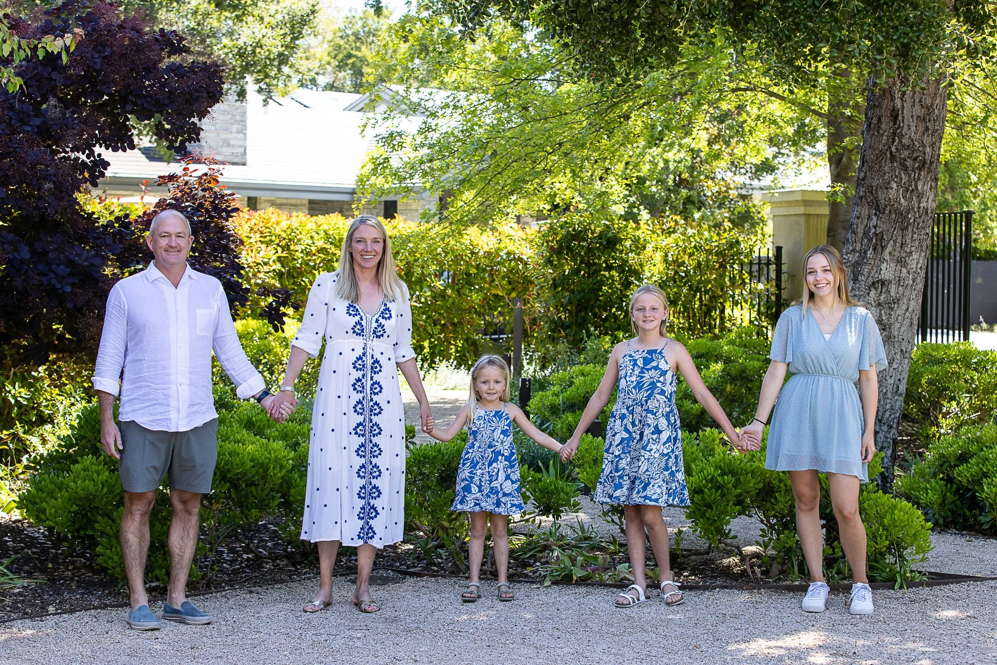 A family of six standing outside on a gravel pathway, holding hands, surrounded by greenery and large trees, with a house partially visible in the background on a sunny day.