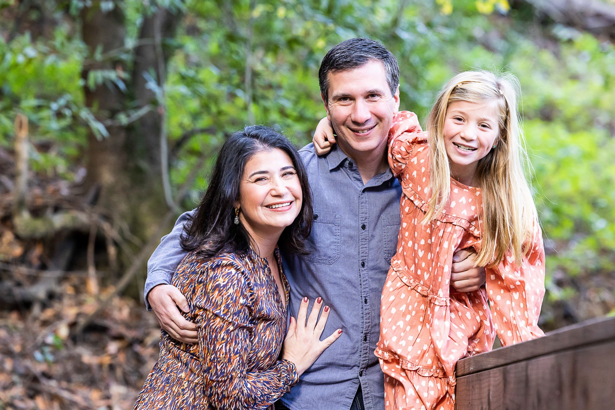 A smiling family of four outdoors in a wooded area, with two women, one man, and a young girl with long blonde hair, all close together and happy.