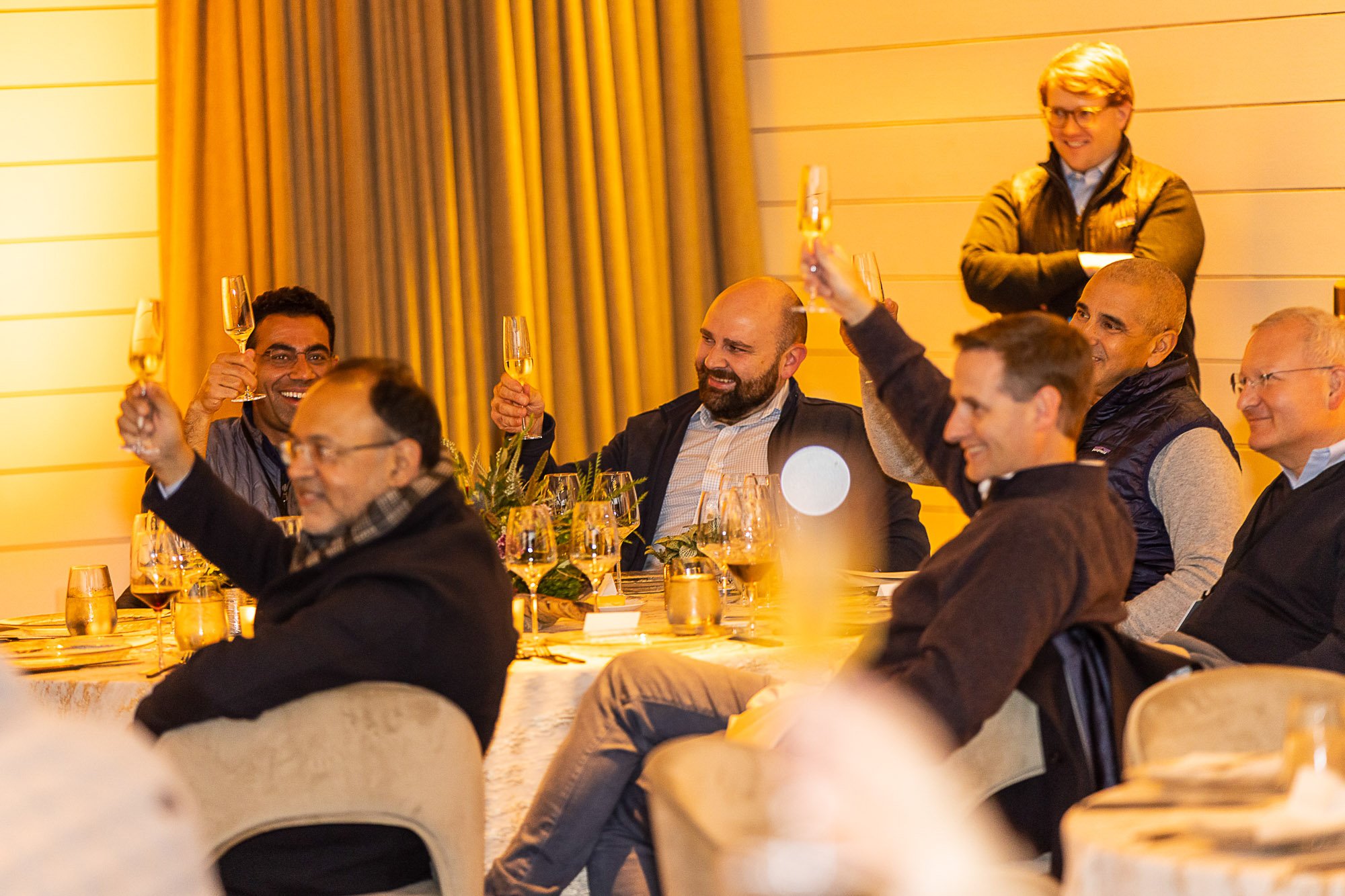 People sitting at a banquet table, raising champagne glasses, smiling, and celebrating in a warmly lit room with yellow walls and curtains.