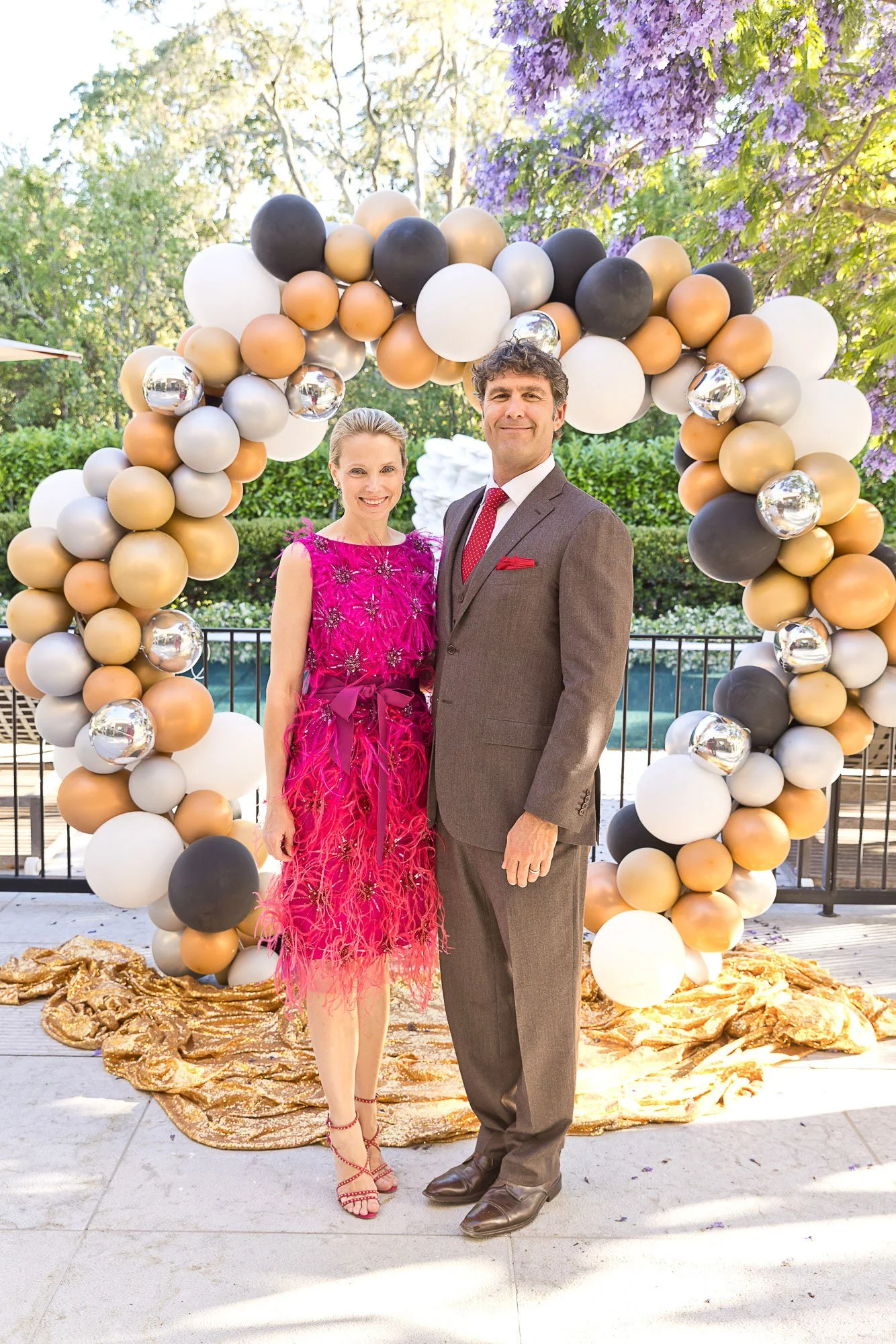 Marissa Mayer in a pink dress and a man in a gray suit stand together, smiling, in front of a decorative balloon arch with black, white, gold, and silver balloons, outdoors with trees and purple flowers in the background.