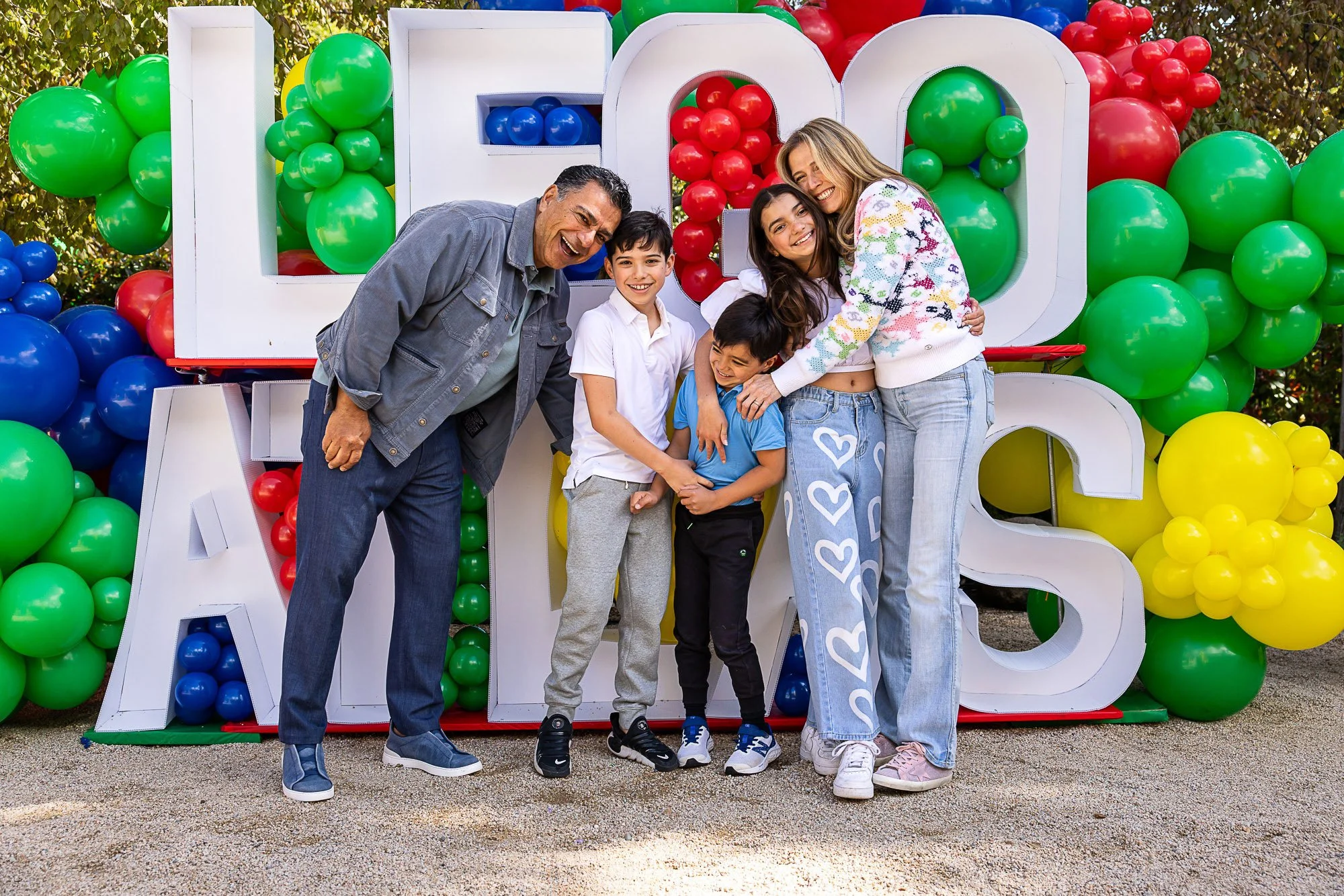 A group of six people, including two adults and four children, smiling and posing for a photo in front of colorful balloon decorations and a large white sign spelling 'LOVE' at an outdoor event.