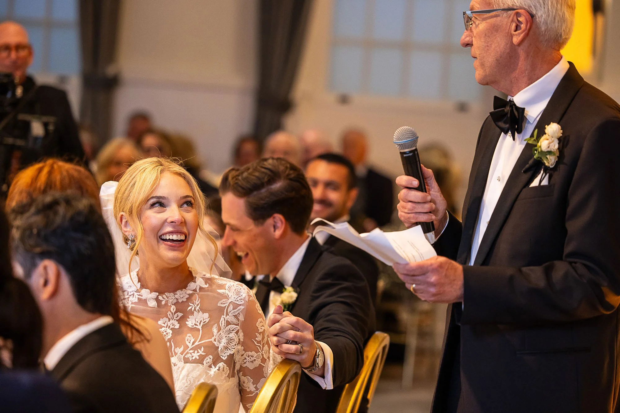 A wedding reception where an elderly man in a tuxedo is giving a speech with a microphone to the bride and groom who are smiling and looking at him. The bride is wearing a lace wedding dress at Cavallo Point in Sausalito, CA.
