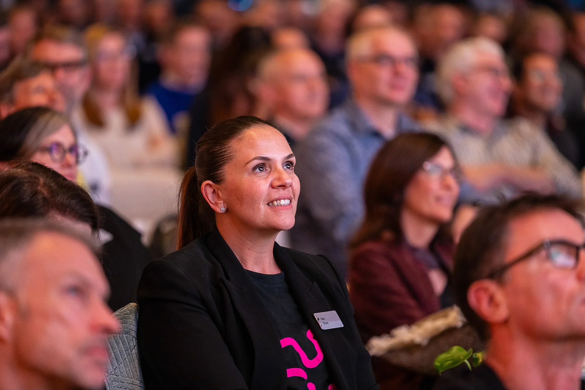 A woman with dark hair pulled back, wearing a black blazer and a name tag, sitting in an audience of mostly women, all attentively listening at an indoor event.