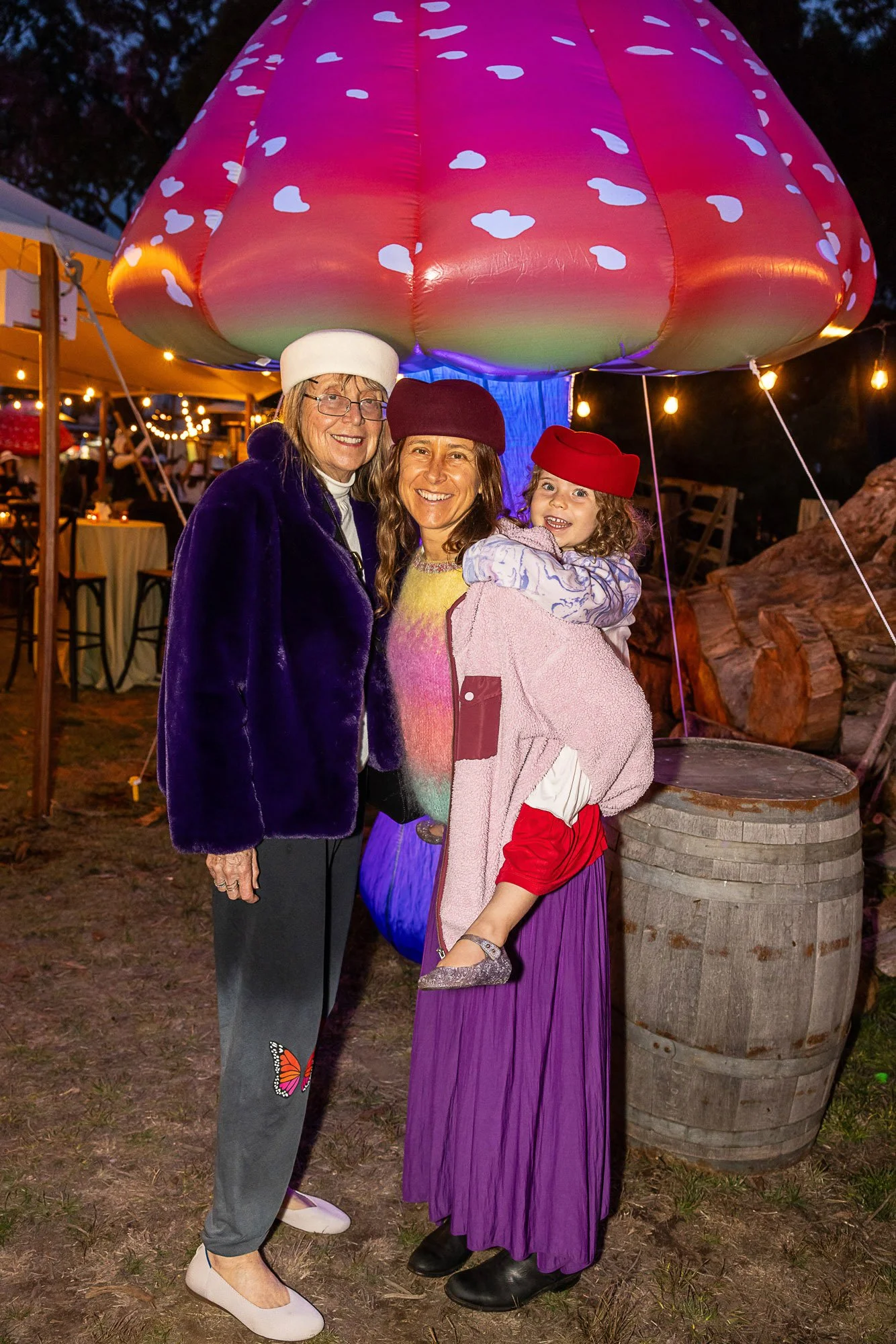 Three people wearing colorful costumes standing under a large, inflatable pink mushroom with white spots, illuminated at night. They are smiling, and there are festively lit tents and outdoor decorations in the background.