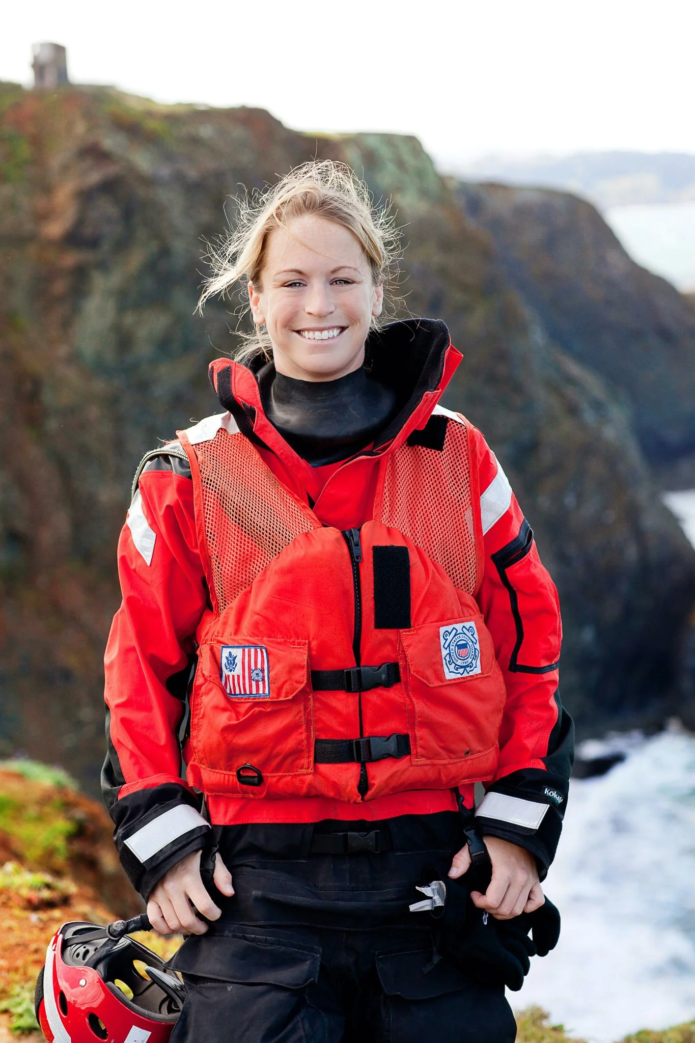 Smiling woman wearing red rescue gear holding a helmet outdoors with a rocky cliff and ocean in the background.
