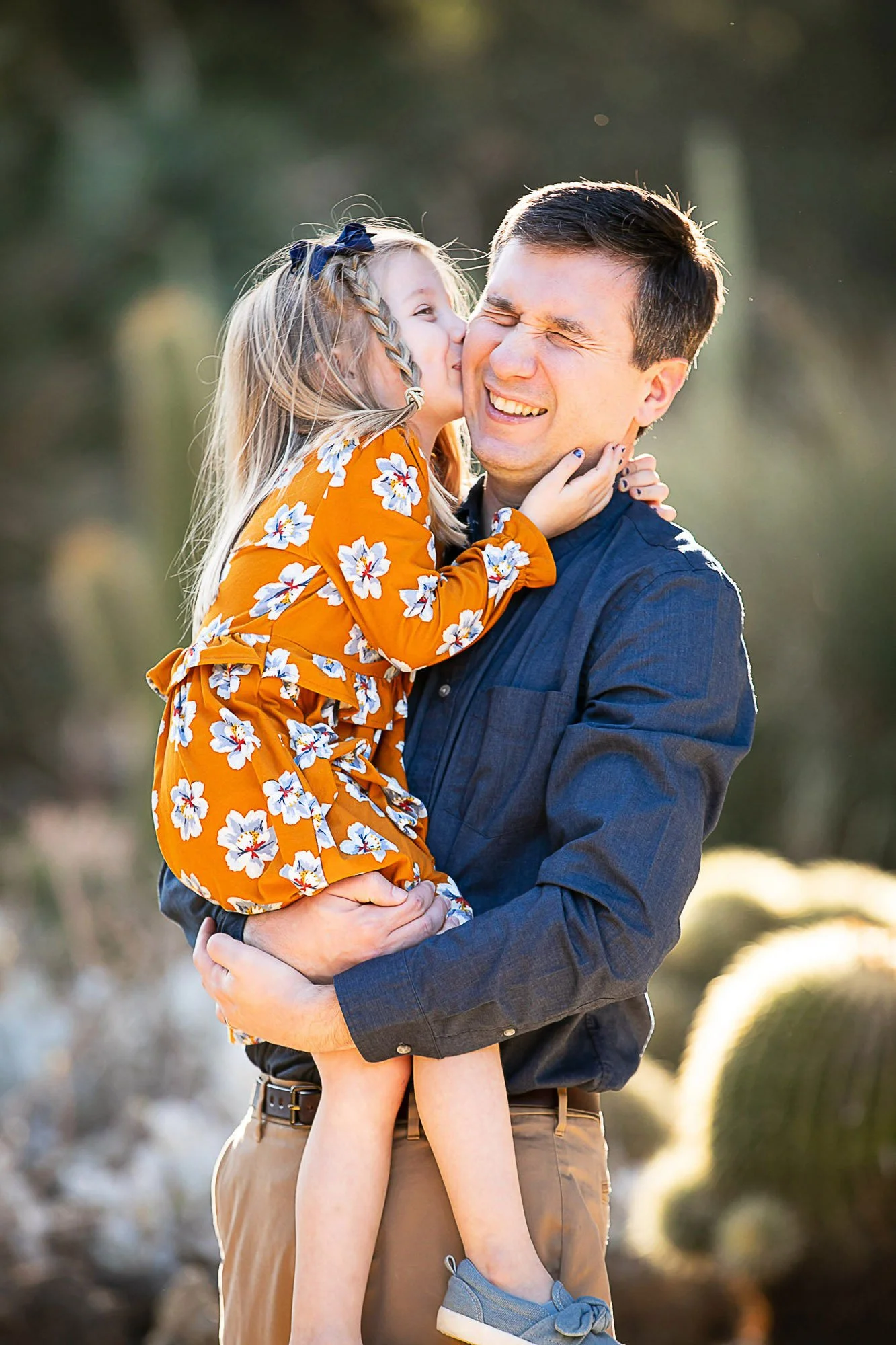 A man holding a young girl who is kissing him on her cheek outdoors during golden hour, with sunlight creating a warm glow.