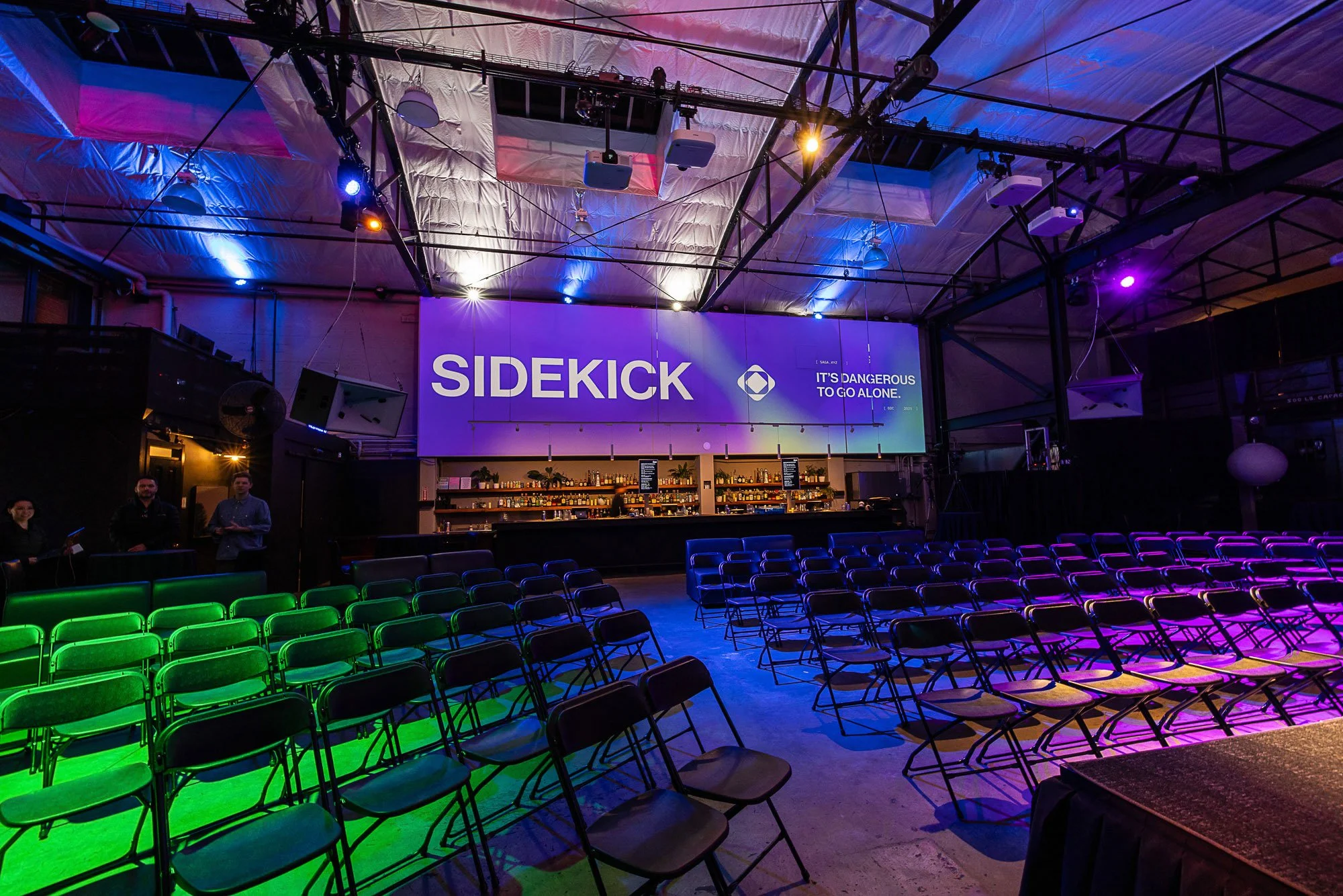 An indoor event venue with rows of green and black chairs facing a stage with a large screen displaying 'SIDEKICK' and the phrase 'It's dangerous to go alone.' The ceiling has colorful lighting, including blue, purple, and orange lights, creating a vibrant atmosphere.