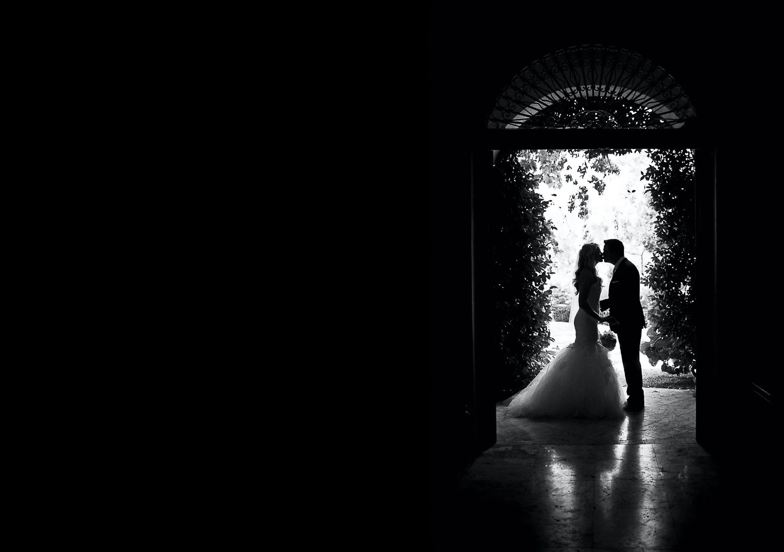 A silhouette of a bride and groom holding hands and touching foreheads in a doorway with intricate metalwork. Bright outdoor light behind them highlights the scene in black and white.