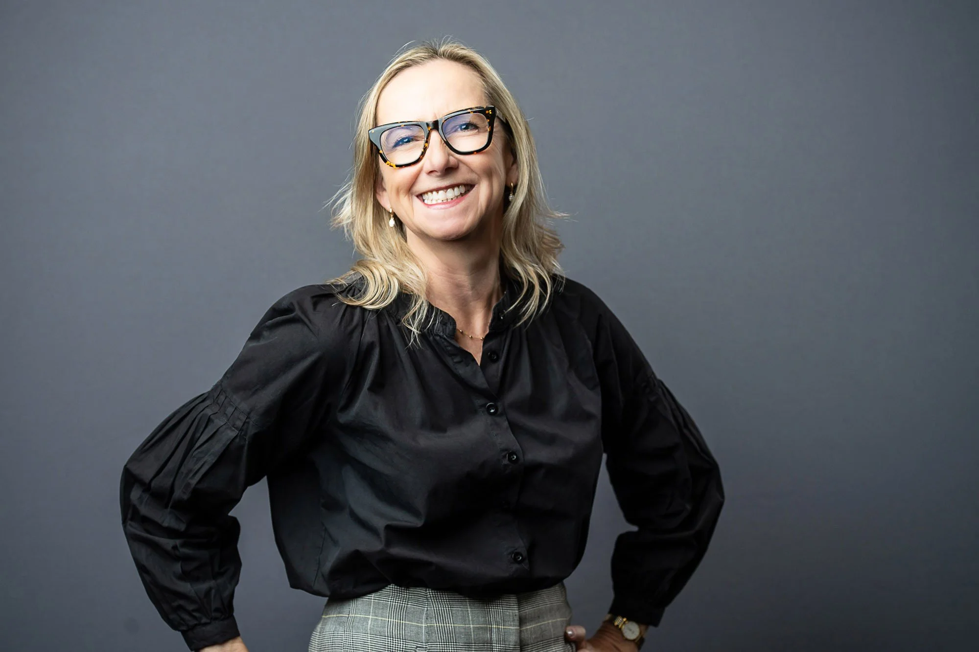 Tara Arrowood with shoulder-length blonde hair, wearing glasses, a black blouse, plaid pants, and accessorized with earrings and a watch, smiling against a gray background.