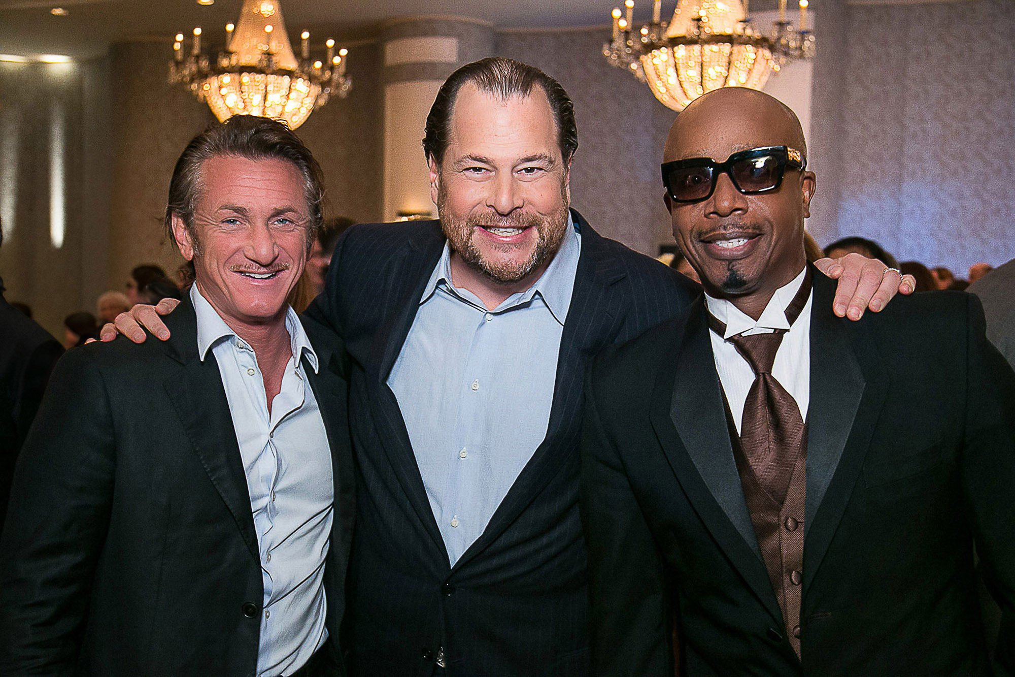 Sean Penn and Marc Benioff, smiling and posing together in an indoor setting with chandeliers and other people in the background at the Fairmont Hotel.