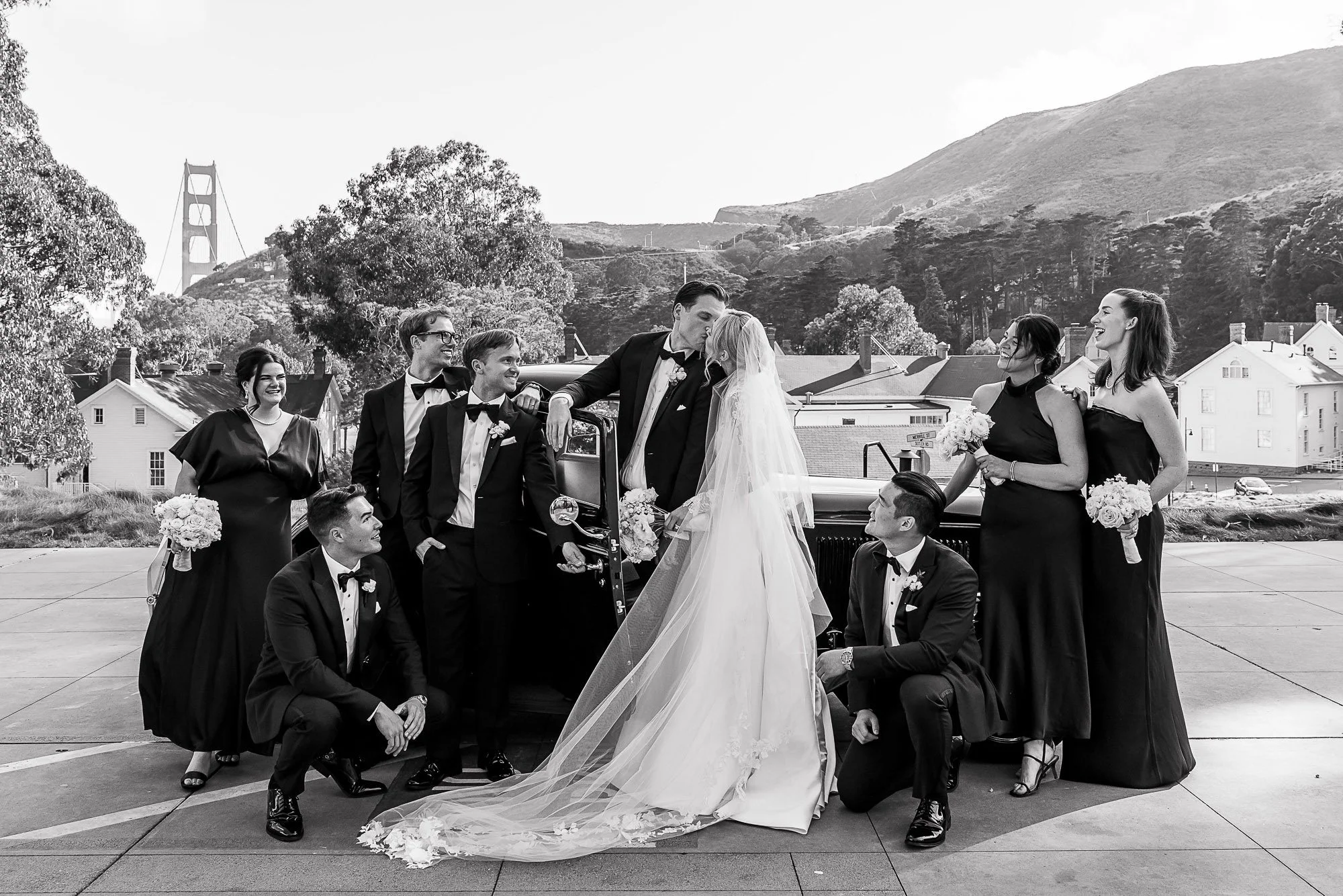 Black and white photo of a wedding party gathering outside with the Golden Gate Bridge in the background. The bride and groom are sharing a kiss while the wedding party surrounds them at Cavallo Point in Sausalito, CA.