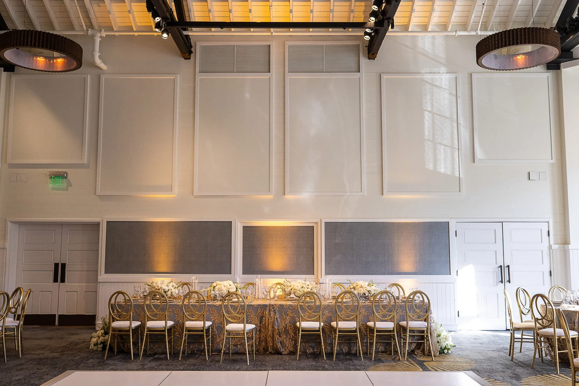 Wedding reception  at Cavallo Point in Sausalito, CA with flowers and gold chairs, set against a wall with square grey panels and white frames, illuminated by warm lighting, in a bright room with a wooden ceiling and large windows.