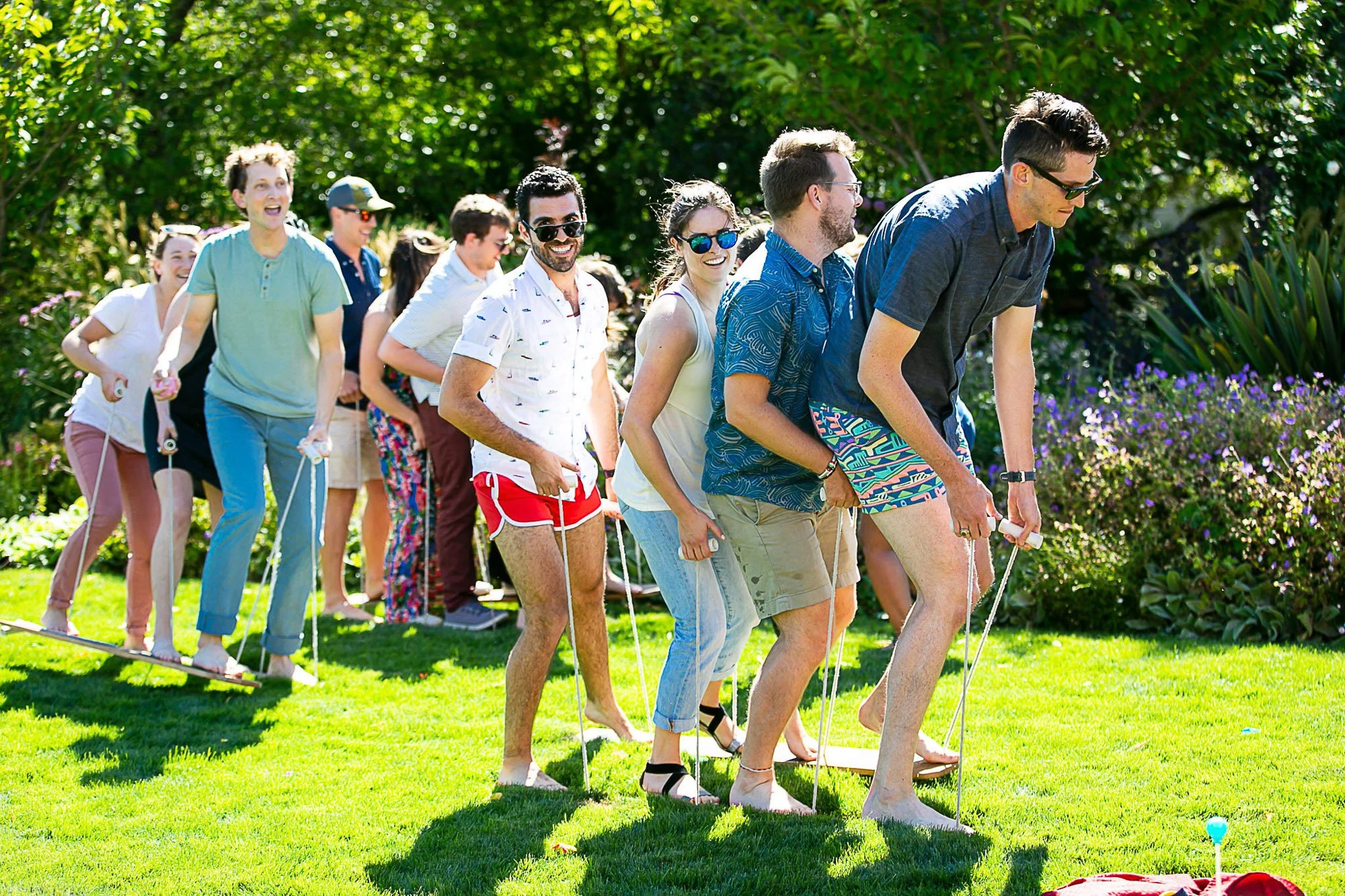 Group of friends playing limbo game outdoors on a sunny day in a lush garden.