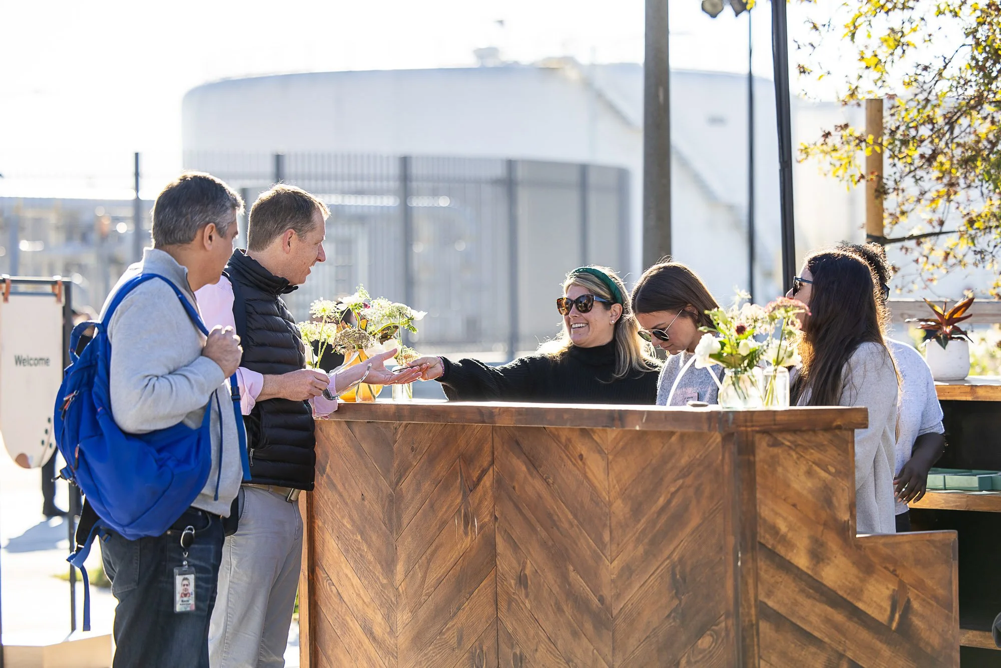 People standing at a wooden table with flowers, engaging in conversation outdoors on a sunny day.