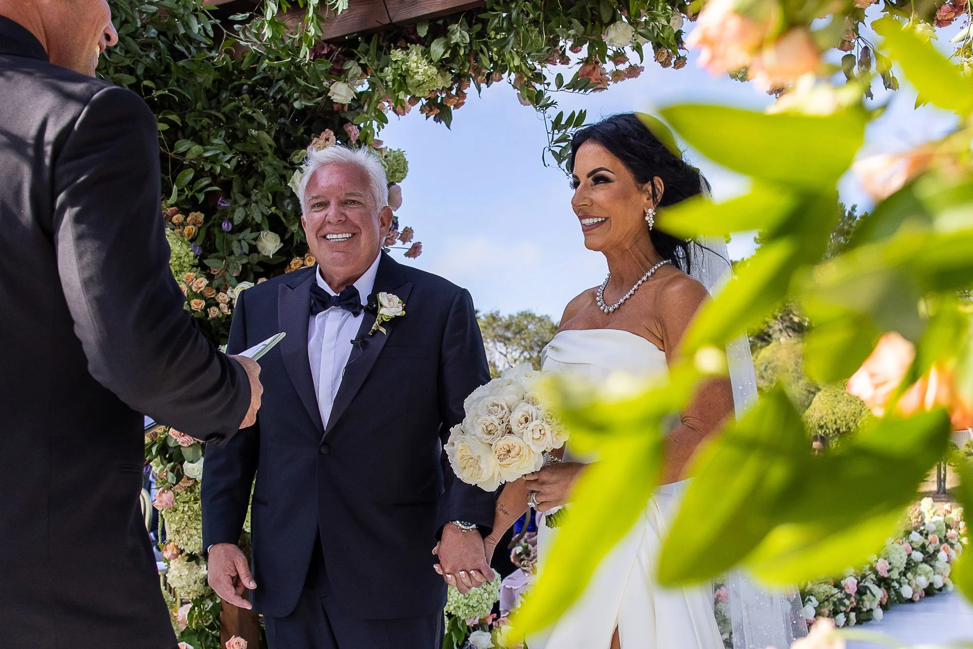 A bride and groom holding hands during their wedding ceremony at the Beach and Tennis Club in Pebble Beach, smiling at each other outdoors, with a floral arch and greenery in the background.
