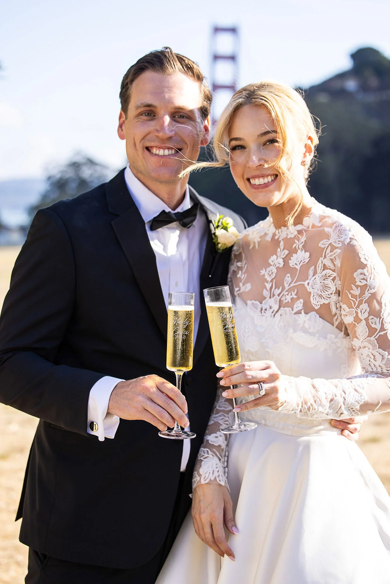 A smiling bride and groom in wedding attire holding glasses of champagne outdoors with the Golden Gate Bridge in the background at Cavallo Point in Sausalito, CA.