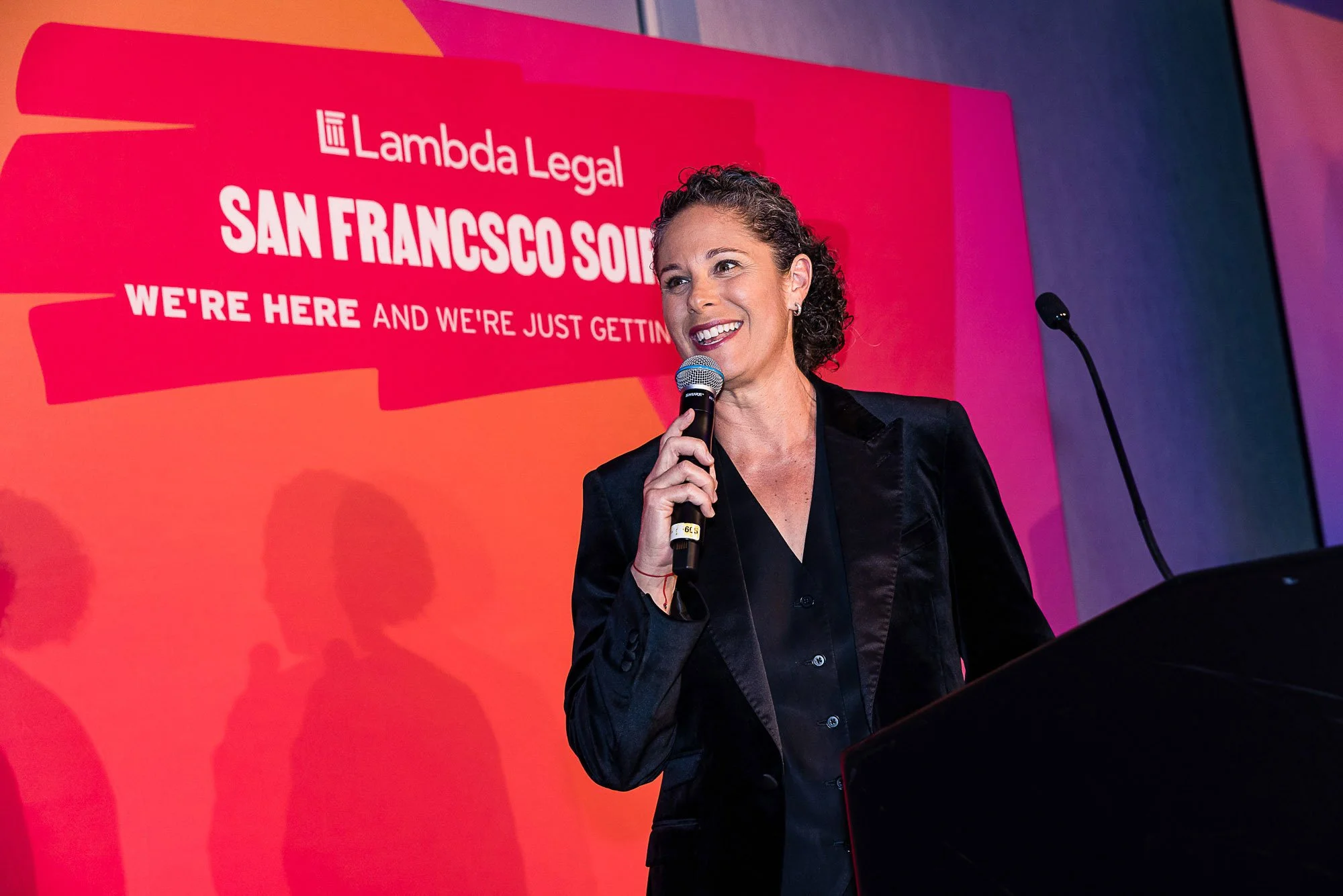 A woman with curly hair and earrings, smiling while speaking into a microphone, at an event with a bright pink backdrop that reads 'Lambda Legal SAN FRANCISCO' and 'WE'RE HERE AND WE'RE JUST GETTING STARTED'.