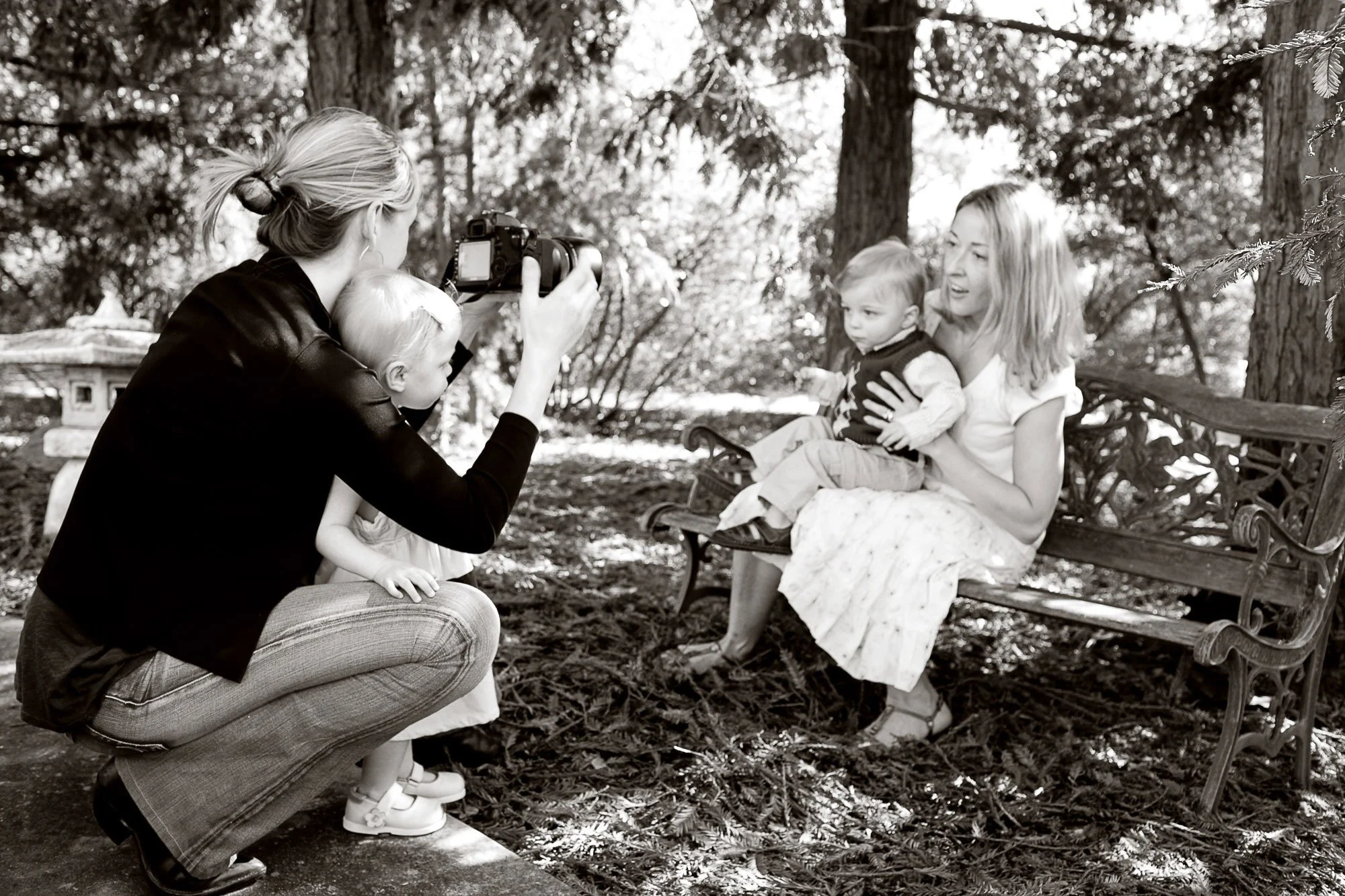 A woman with long hair sitting on a park bench holding a small child on her lap while another woman kneels in front of them taking a photo with a camera. A woman with a bun hairstyle is also present, leaning in towards the woman on the bench. The scene is set outdoors with trees in the background.