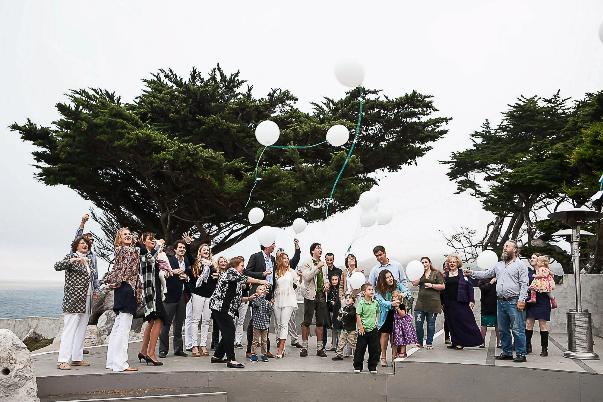 A group of people gathered outdoors near trees and a body of water, releasing white balloons into the sky during an overcast day.