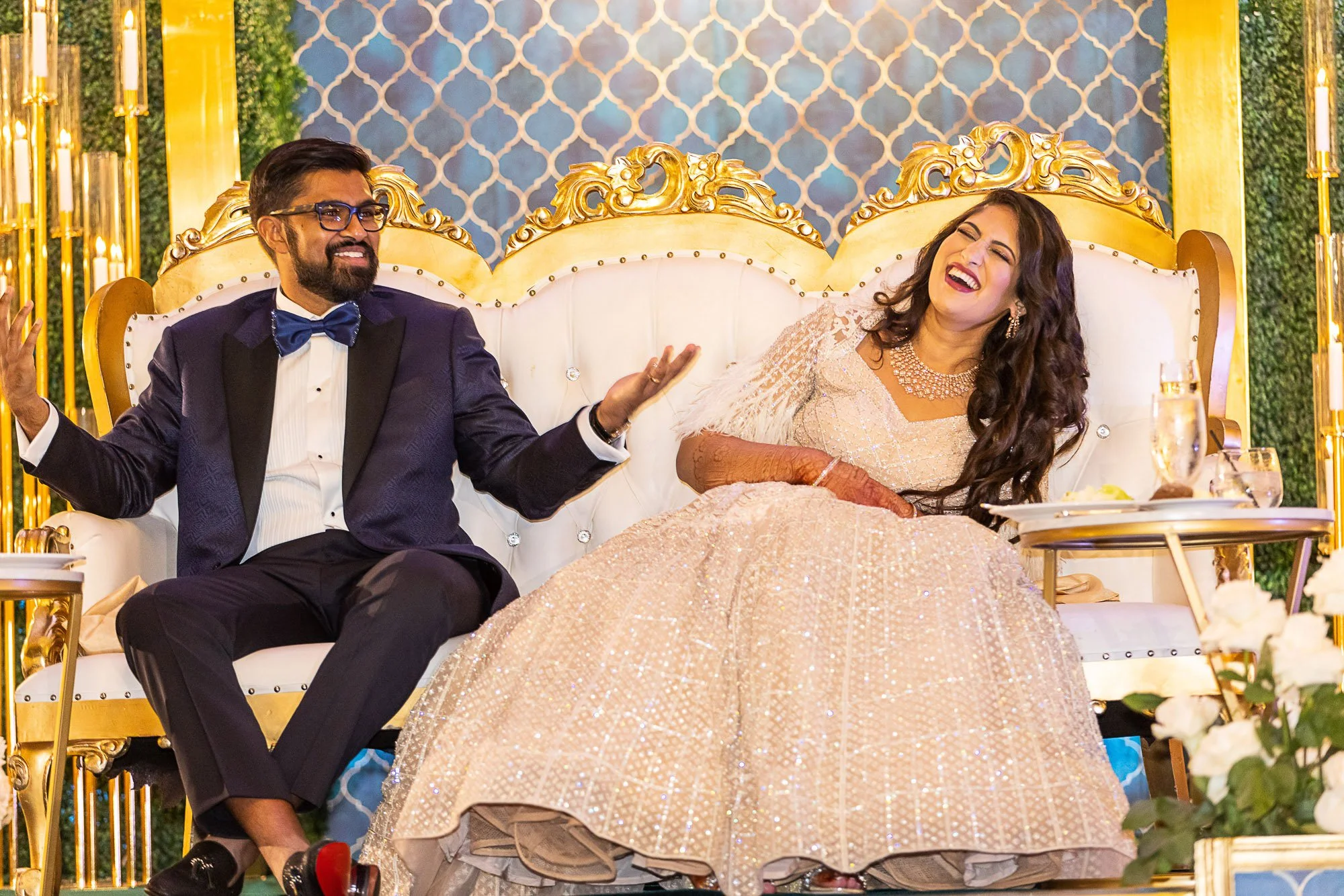 A man in a tuxedo and a woman in a shimmering gown are sitting on a fancy white sofa with gold ornate detailing. They are laughing and appear to be enjoying a wedding reception at the Palace Hotel San Francisco with candles and floral arrangements.