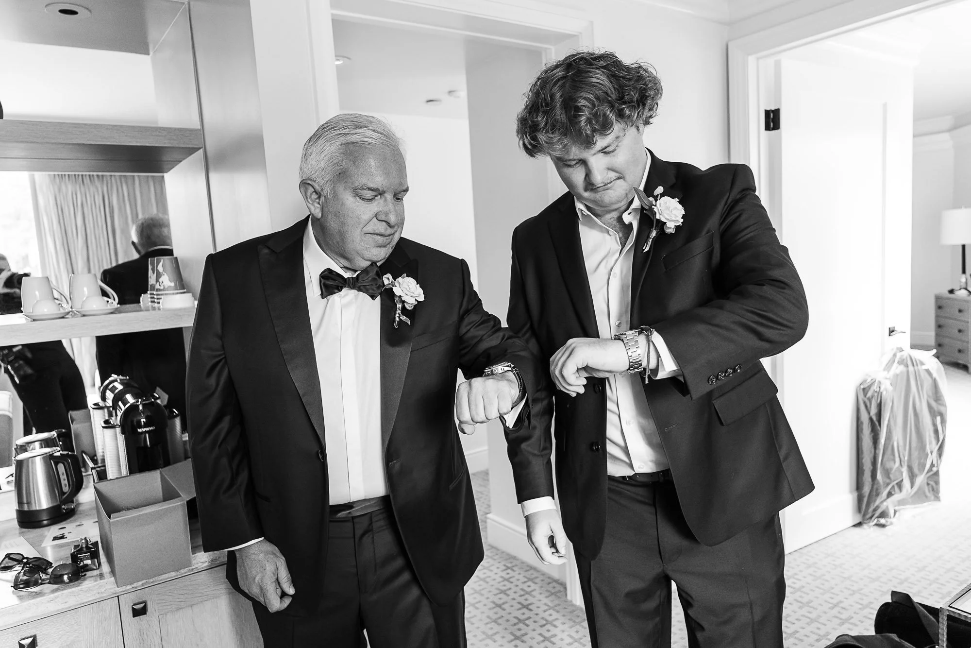 Two men in formal suits at the Beach and Tennis Club in Pebble Beach, CA, one elderly and one younger, looking at their watches in a hotel room, with a mirror, coffee cups, and luggage visible in the background.