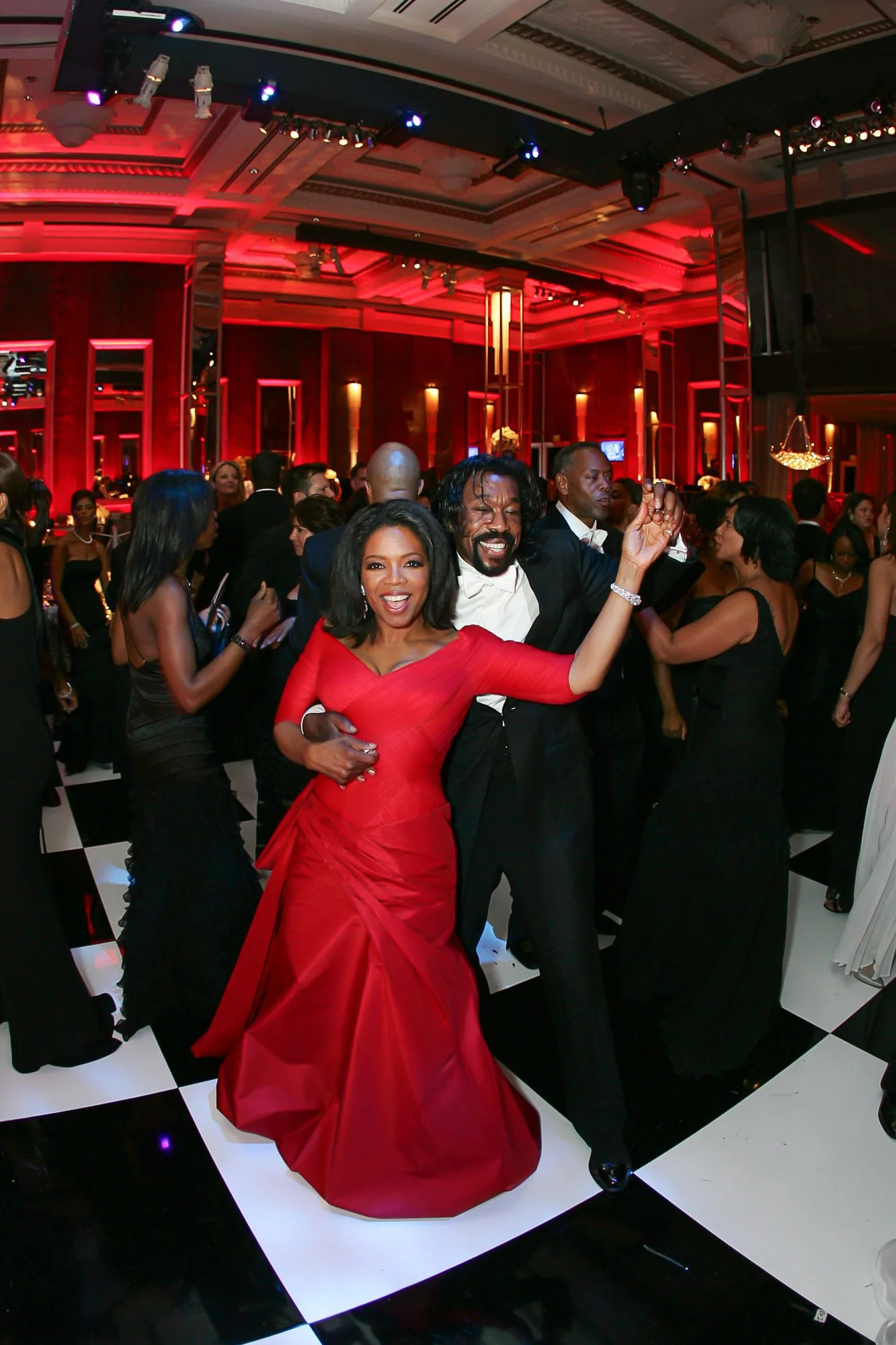 Oprah Winfrey dancing at a formal event in a grand hall with red lighting. Prominent figures, including a woman in a red dress and a man in a tuxedo, are smiling and enjoying themselves.